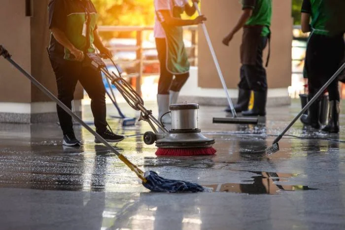 People cleaning a wet outdoor floor with brushes, mops, and a floor scrubber machine in sunlight.