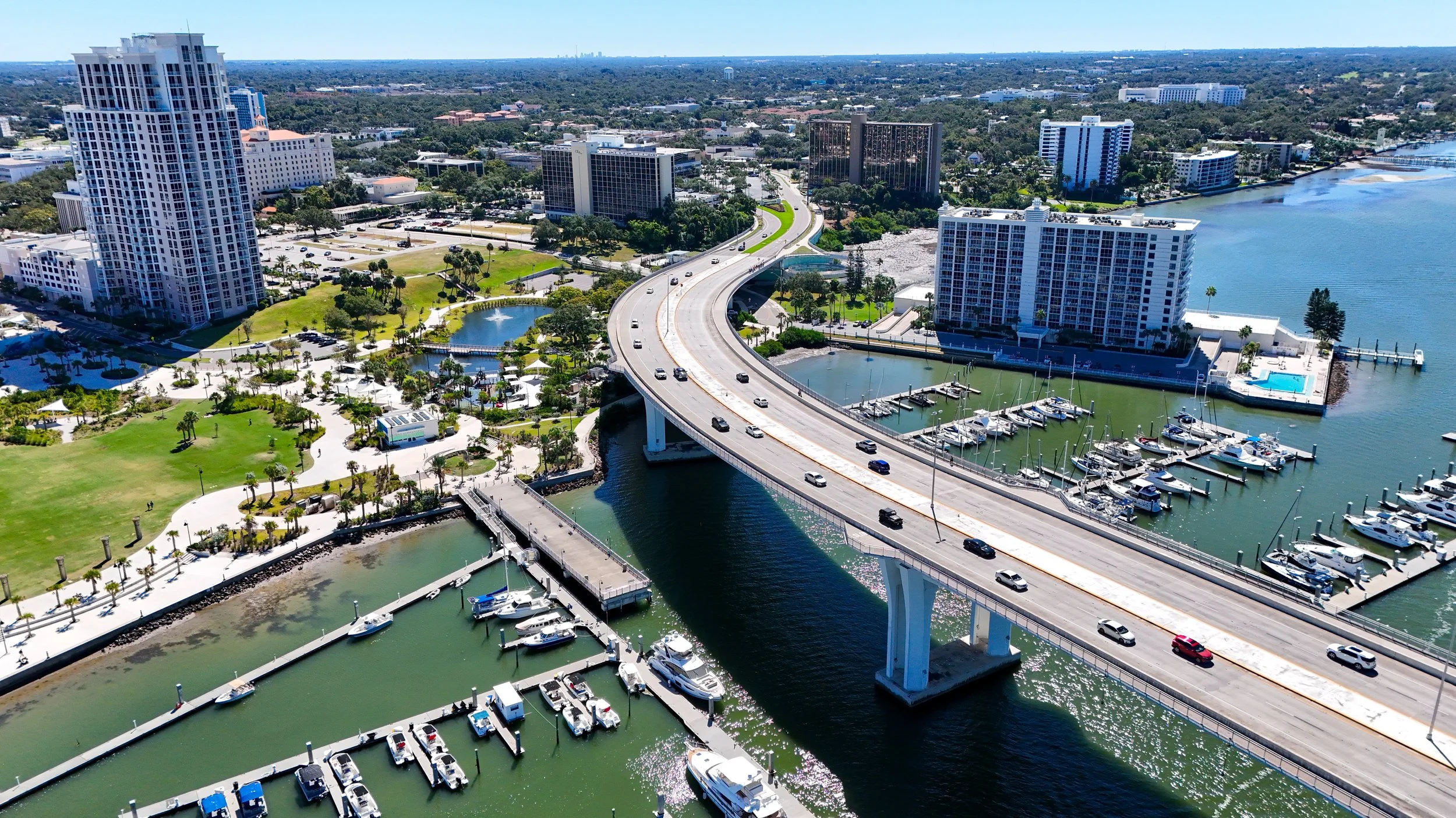 Aerial view of a city with a bridge crossing over a waterway, surrounded by high-rise buildings, marina with boats, and green parks.