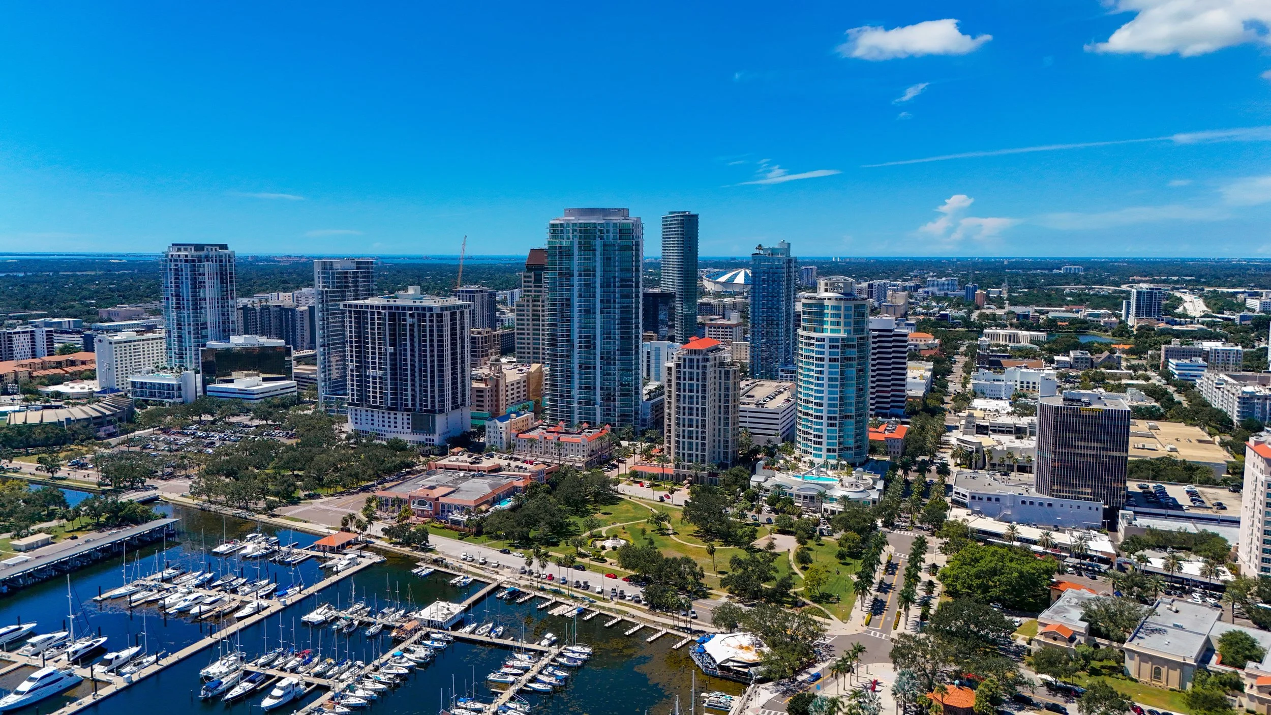 Aerial view of a city skyline with tall modern buildings, marina with boats, and green parks under a partly cloudy blue sky.