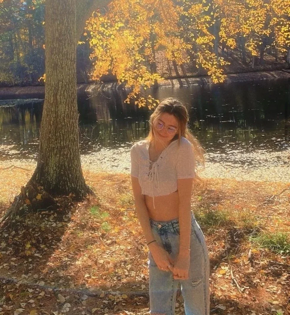 A teenage girl with glasses and long hair standing outdoors by a tree near a lake during autumn in the daytime. Sunlight filters through yellow leaves.