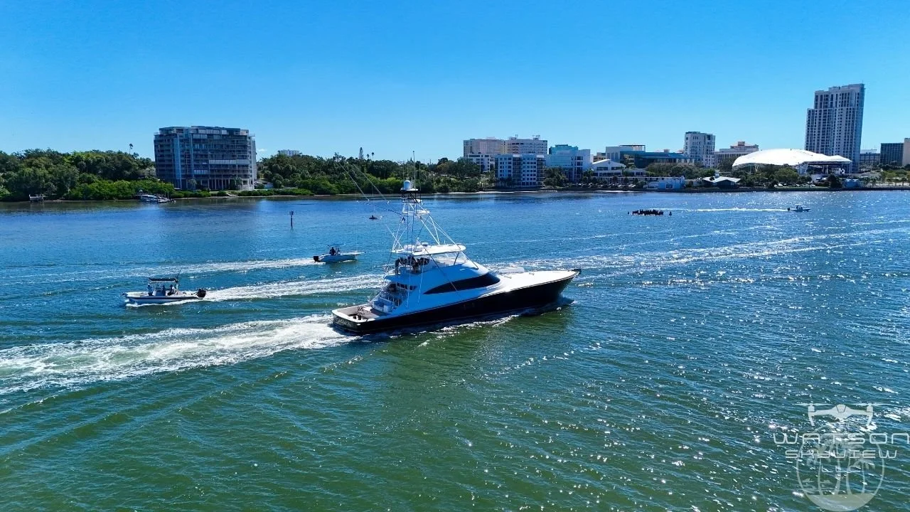 A large yacht cruising on a river near a city, with multiple smaller boats around it and a city skyline with tall buildings in the background.