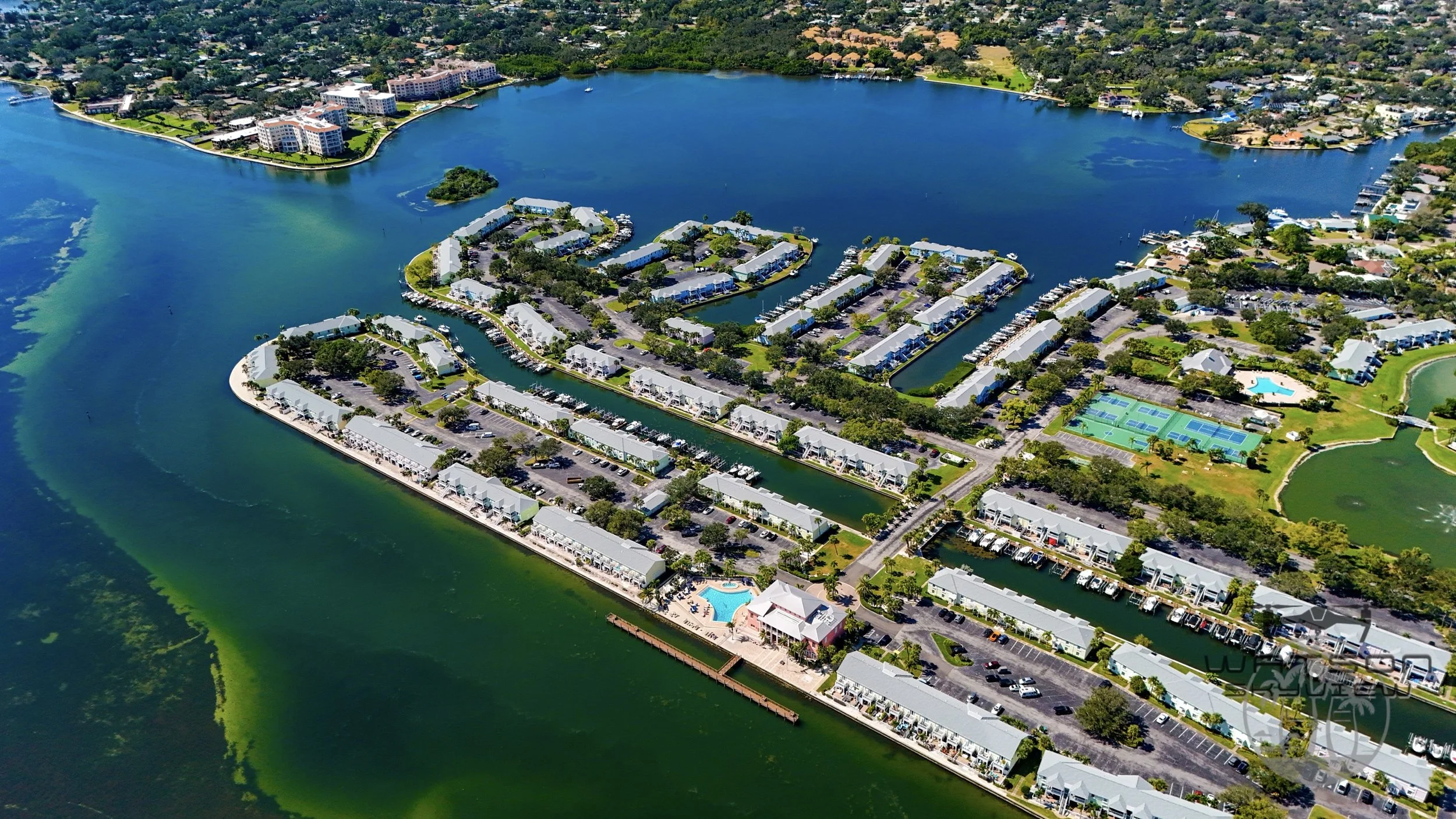 Aerial view of a residential water community with houses, parking lots, a pool, and tennis courts surrounded by a large lake, with greenery and additional buildings in the distance.