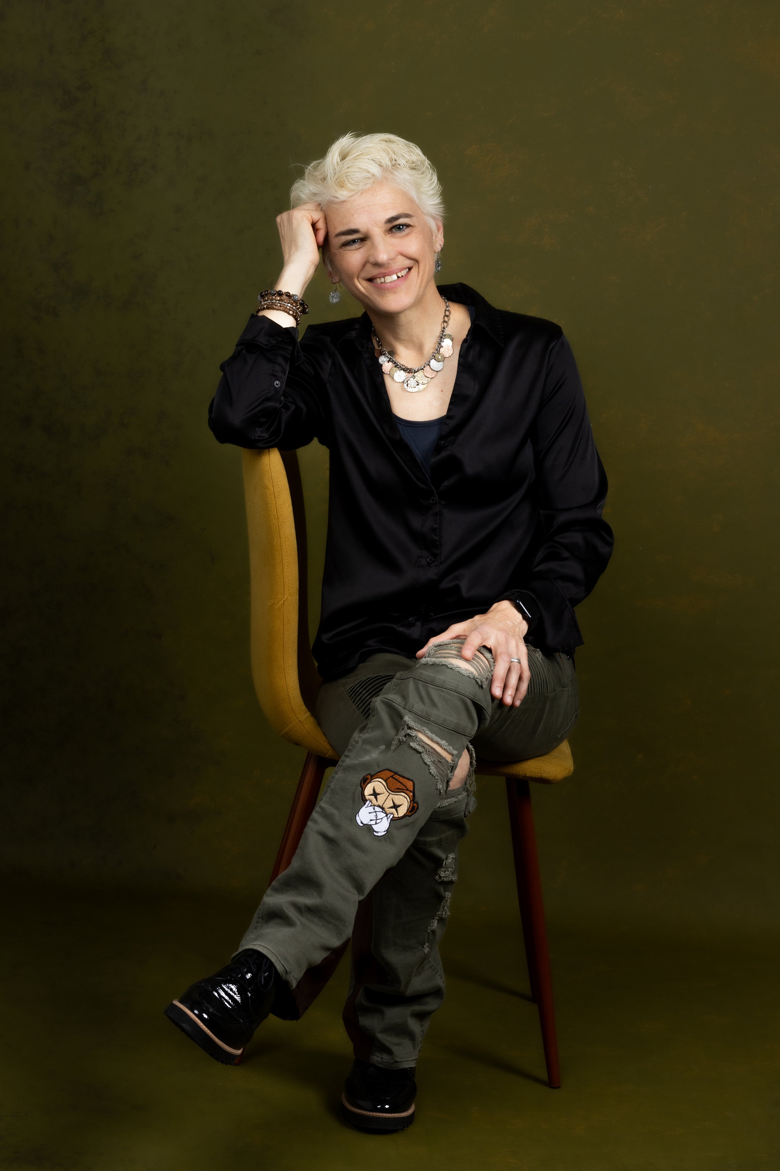 A woman with short, curly white hair smiling while sitting on a yellow chair against a dark green background. She is wearing a black satin shirt, layered necklaces, bracelets, ripped cargo pants with a patch, and black shoes.
