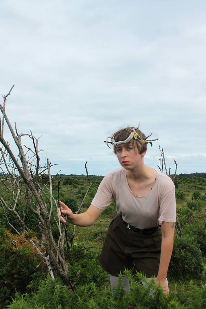 A young woman with short, light brown hair, wearing a beige T-shirt and brown shorts, standing in a field of green and purple bushes under a cloudy sky. She is holding a stick and wearing a crown made of twisted branches.