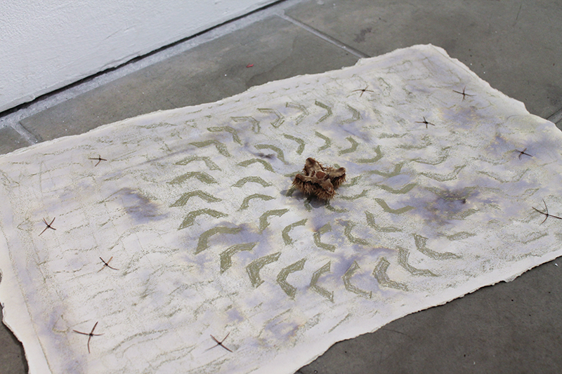 A textured white fabric with a small plush bear in the center, decorated with zigzag lines and crossed marks, laid on a concrete floor next to a white wall.