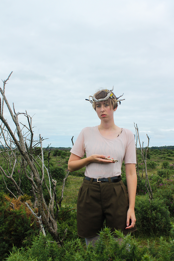 Young woman with a crown made of twigs standing in a green, shrub-filled field on a cloudy day.