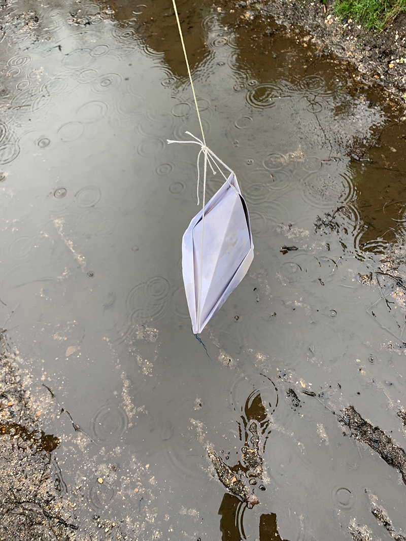 A paper boat hanging from a string above a muddy puddle with raindrops creating ripples.