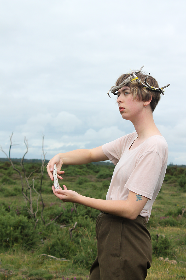 Young woman with a crown made of twisted branches and leaves, holding a piece of driftwood, outdoors in a grassy field with a cloudy sky.