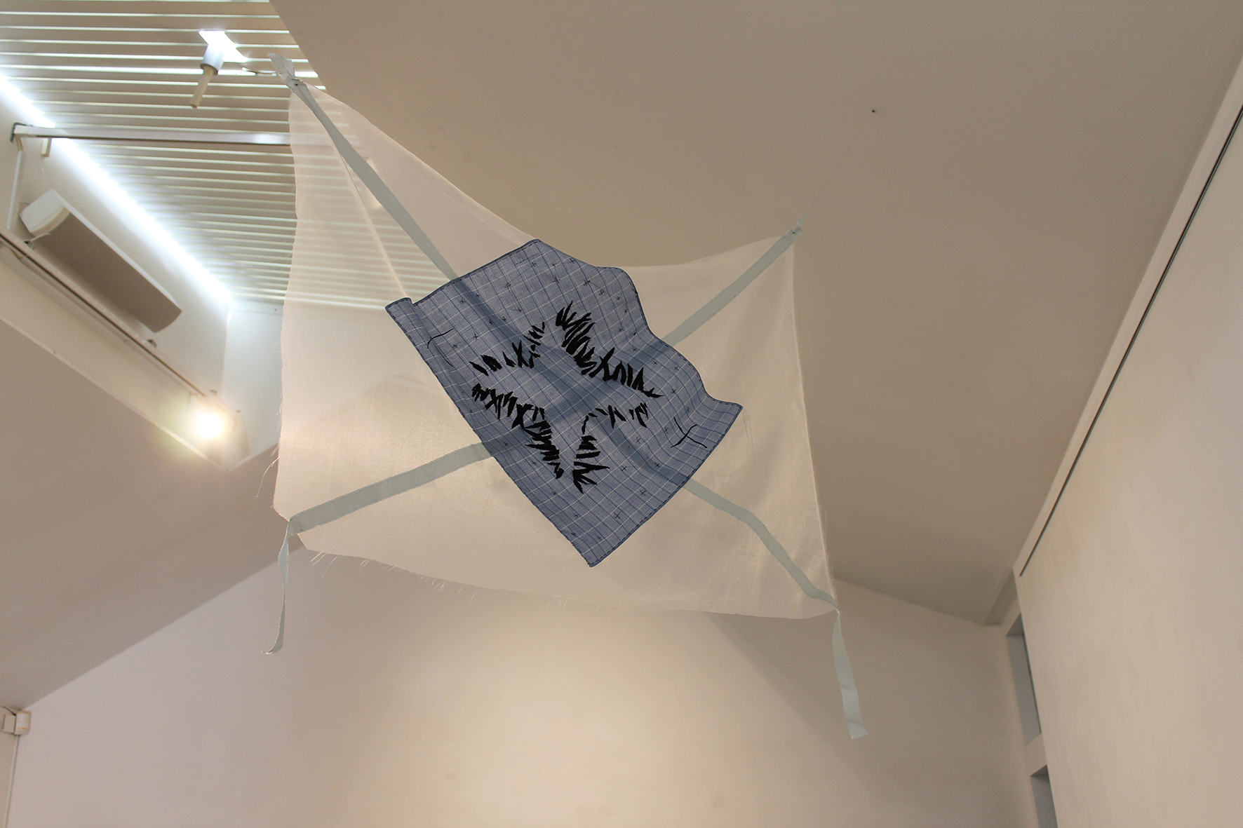 A white fabric object, possibly a kite or decorative banner, hanging from the ceiling with a blue plaid cloth in the center that has black embroidered or printed text. The ceiling is white with a light fixture and an air conditioning unit visible.