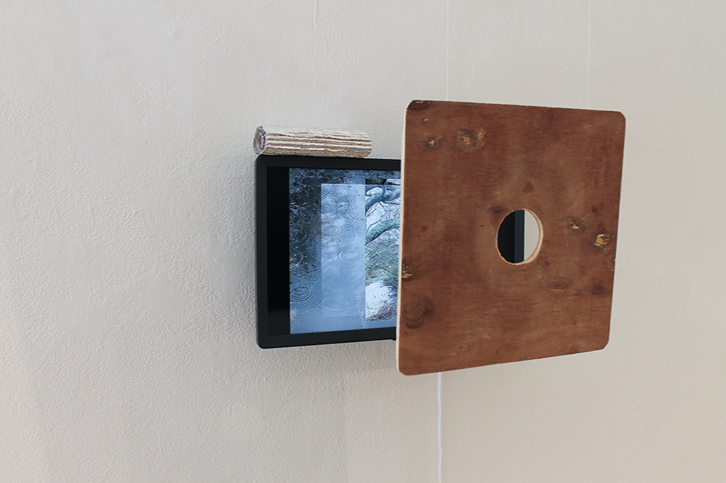 A small black digital photo frame mounted on a wall, partly covered by a square wooden board with a circular hole in the center, with a piece of wood resting on top of the frame.