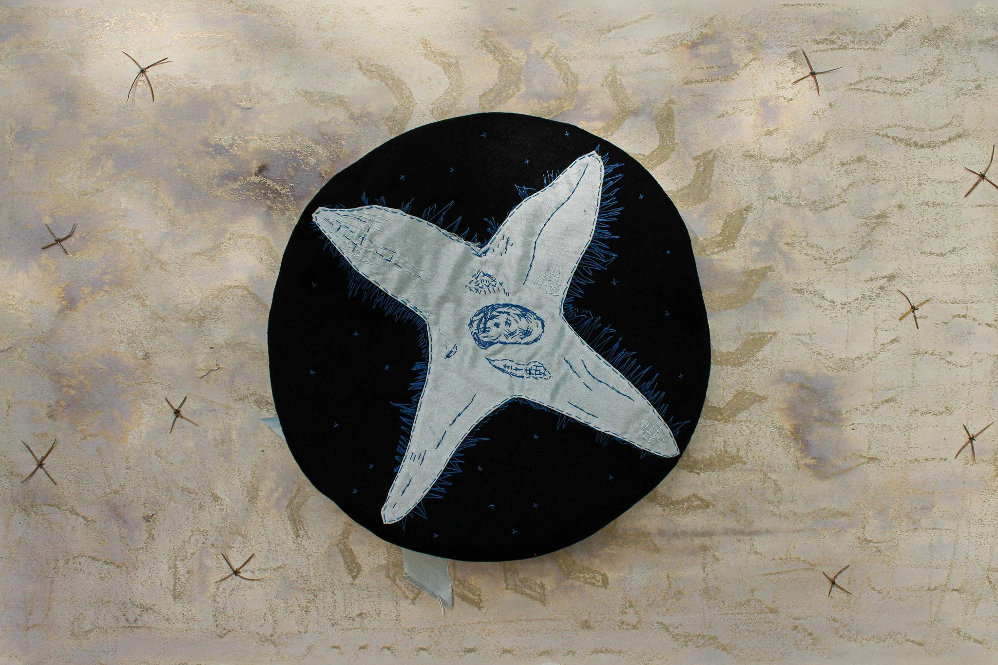 A fabric art piece depicts a white starfish with detailed stitching, placed against a black circular background, embellished with blue thread accents, on a textured beige surface with scattered small, crossed-stitch decorative elements.