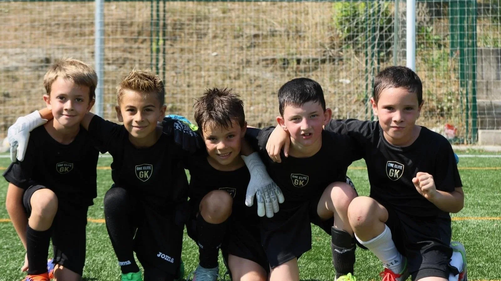 Grupo de cinco niños en uniforme de fútbol, posando en un campo con césped artificial y alambrado de fondo, en actitud de amistad y celebración.
