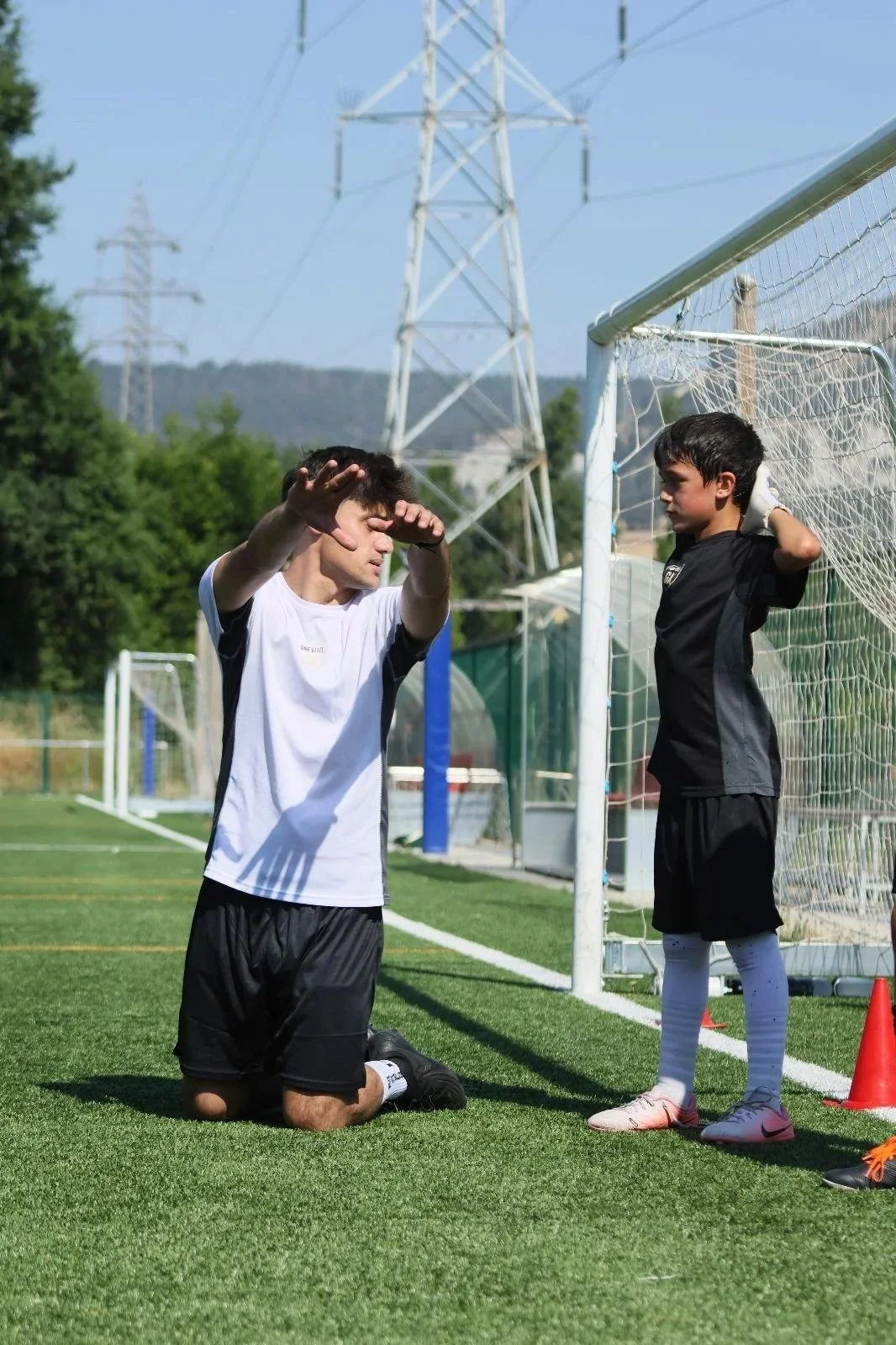 Un niño en posición de rodillas en un campo de fútbol, aparentemente pidiendo ayuda o en una situación de desesperación, mientras otro niño, que parece ser un arquero, se encuentra parado junto a él, apoyado en el arco de fútbol.