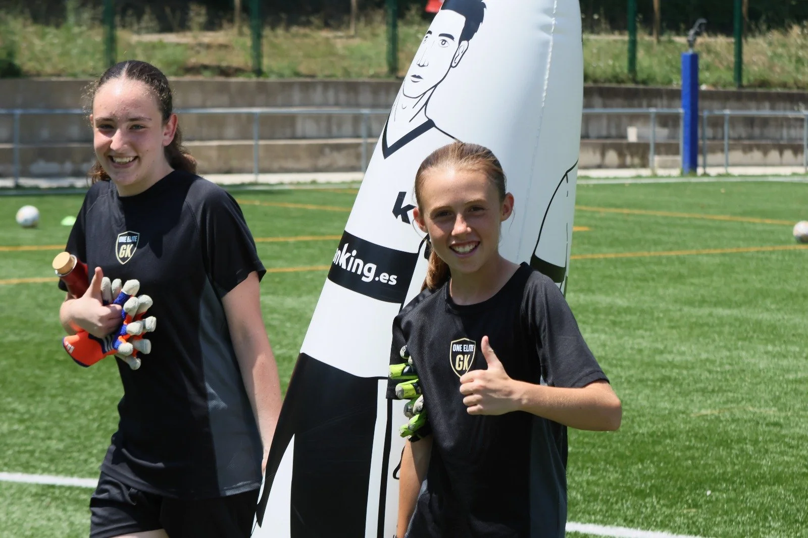 Dos jóvenes futbolistas femeninas en un campo de fútbol, una de ellas sosteniendo guantes y un balón, ambas sonriendo y con uniforme negro.