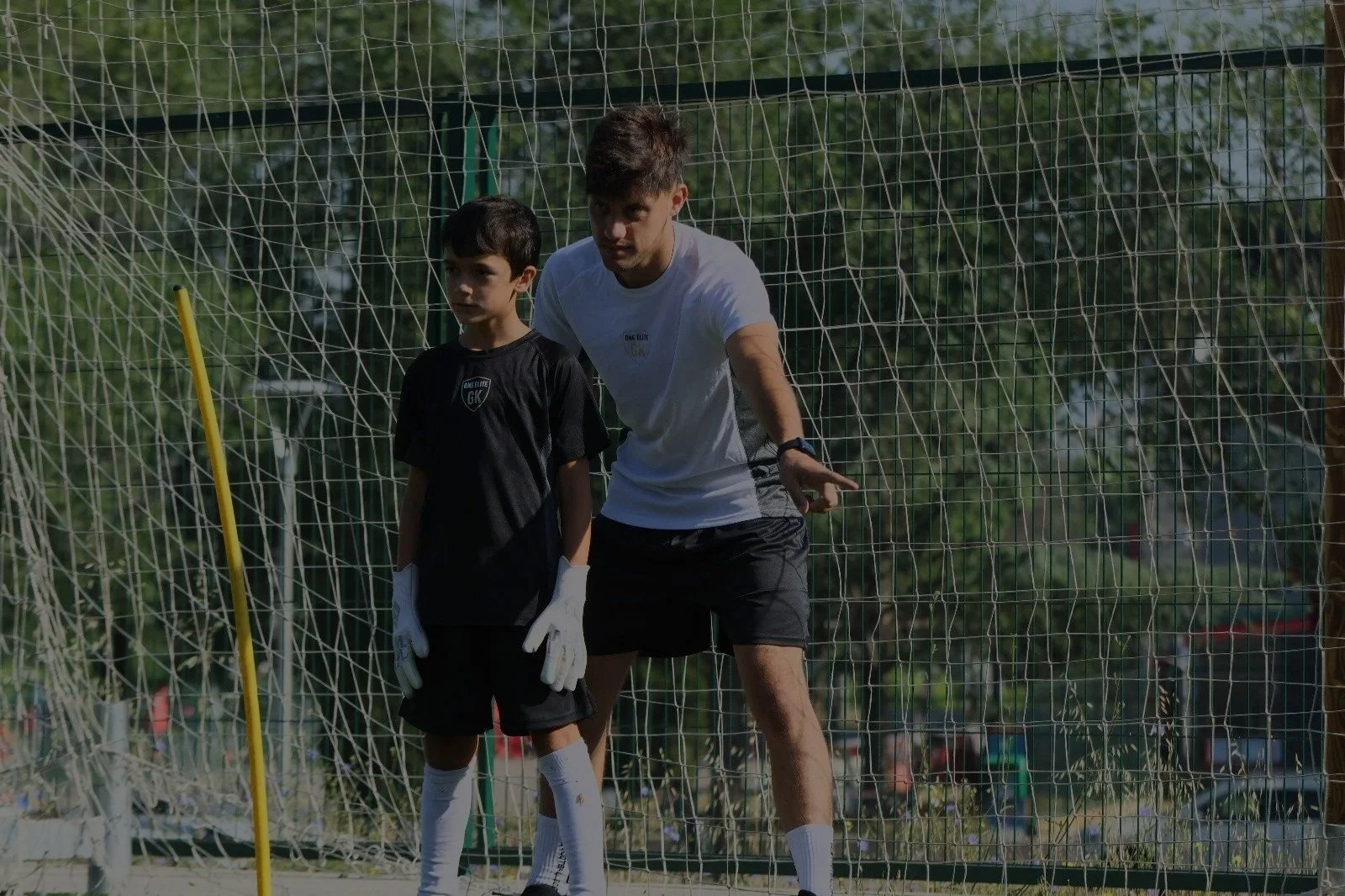 Entrenador entrenando a un niño en una cancha de fútbol, ambas personas están en la portería y están en presencia de una red de fútbol.