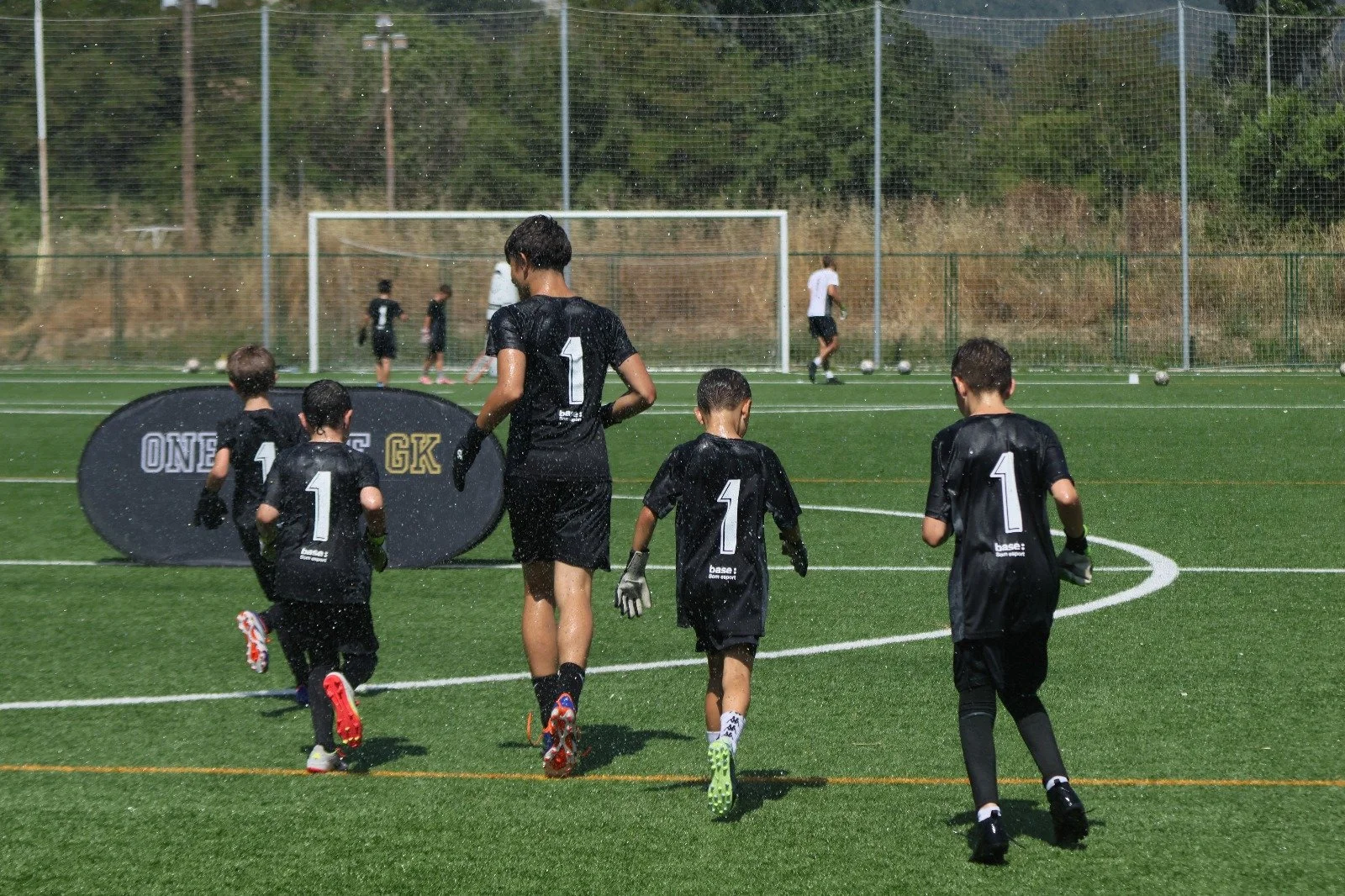 Grupo de niños y un adolescente jugando fútbol en un campo con porterías y una malla de protección, algunos con guantes y camisetas negras con el número uno en la espalda, en un día soleado con árboles al fondo.