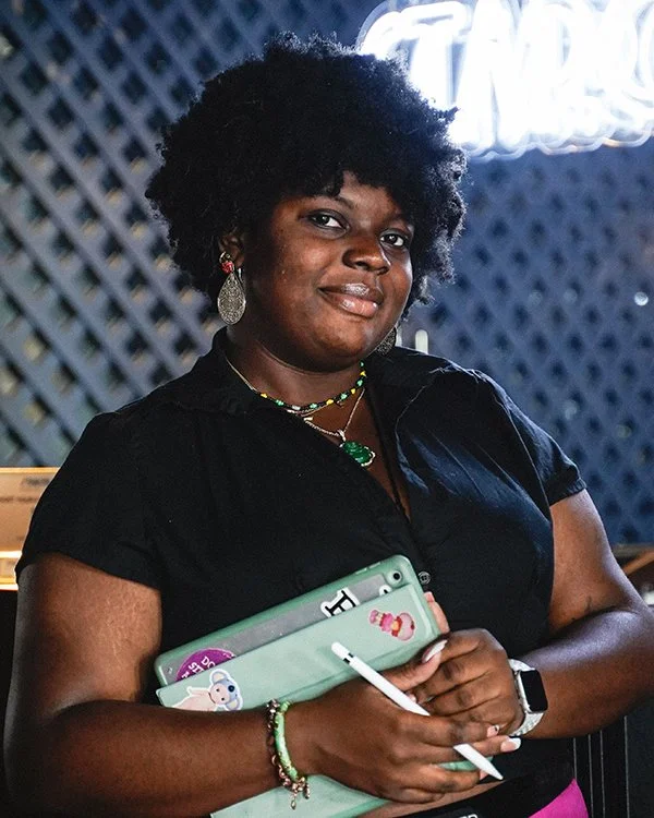 A woman with dark skin, curly black hair, and earrings standing indoors. She is wearing a black shirt, colorful jewelry, and a smartwatch. She holds a light green tablet with stickers on it and a stylus in her hands.