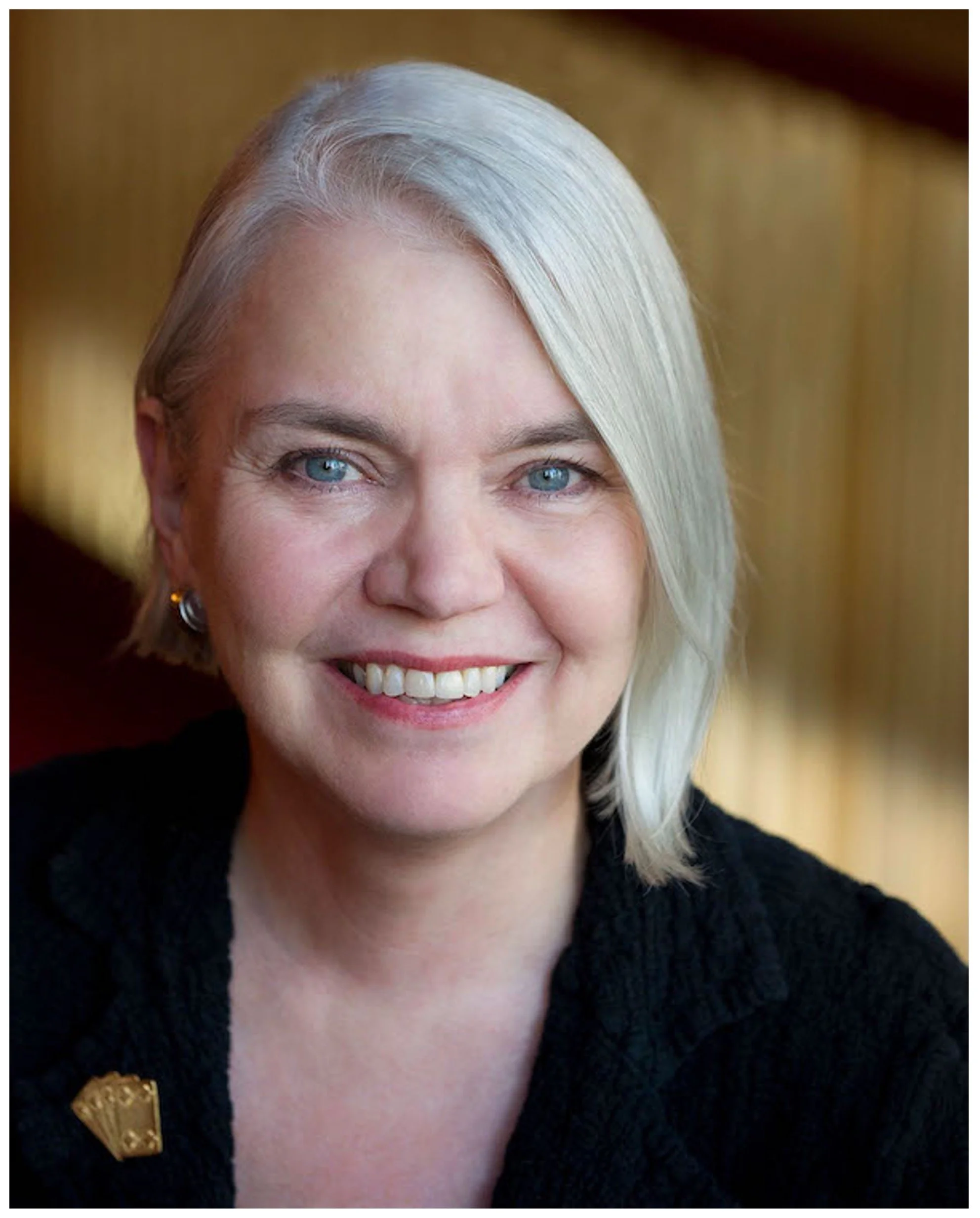 Close-up portrait of a woman with short gray hair, blue eyes, and a bright smile, wearing a black top with a gold brooch, in a warm indoor setting.