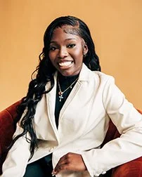 A smiling young woman with long black hair, wearing a white blazer and a gold necklace, sitting on a sofa against a yellow background.