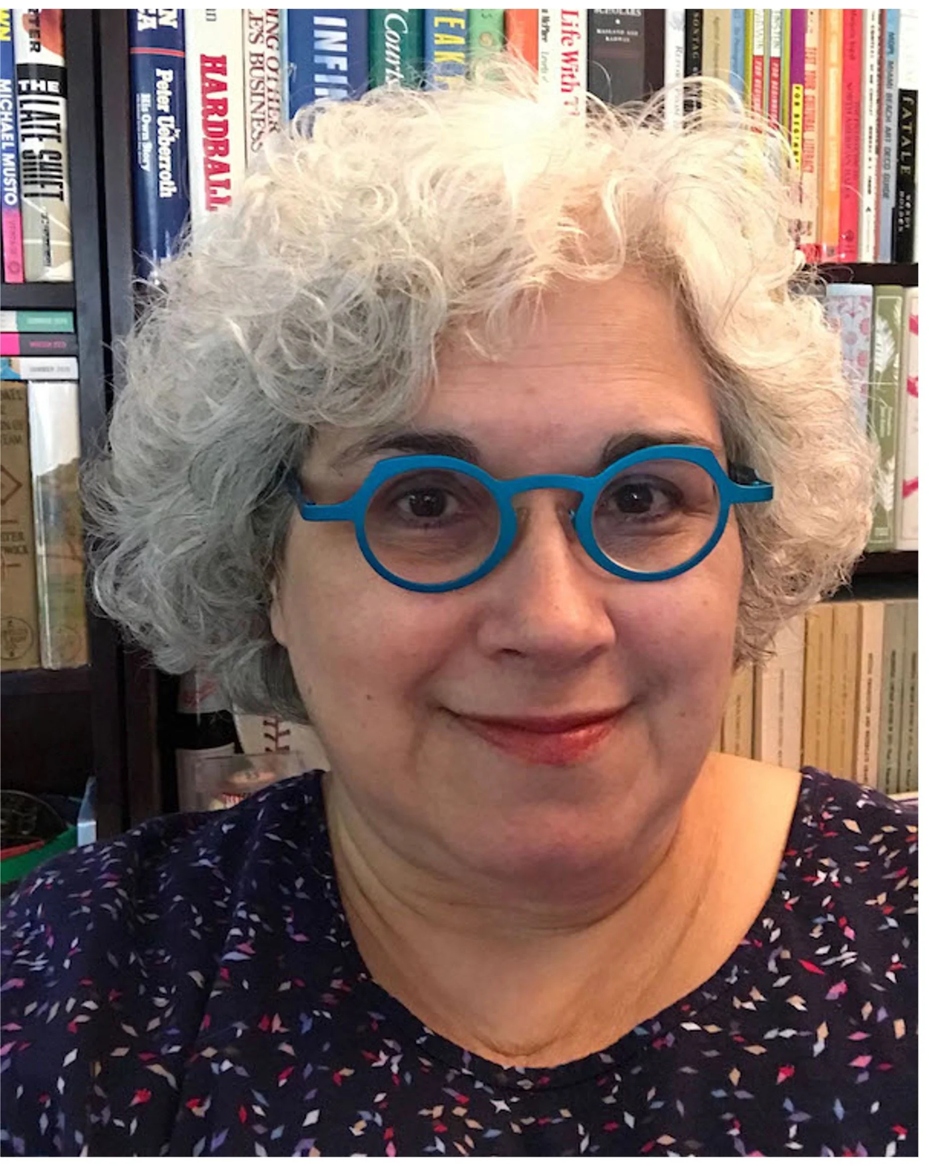 A woman with curly gray hair wearing blue round glasses, standing in front of a bookshelf filled with books.