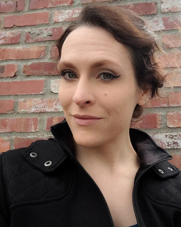 A woman with short brown hair and light makeup standing outdoors in front of a brick wall, wearing a black jacket.