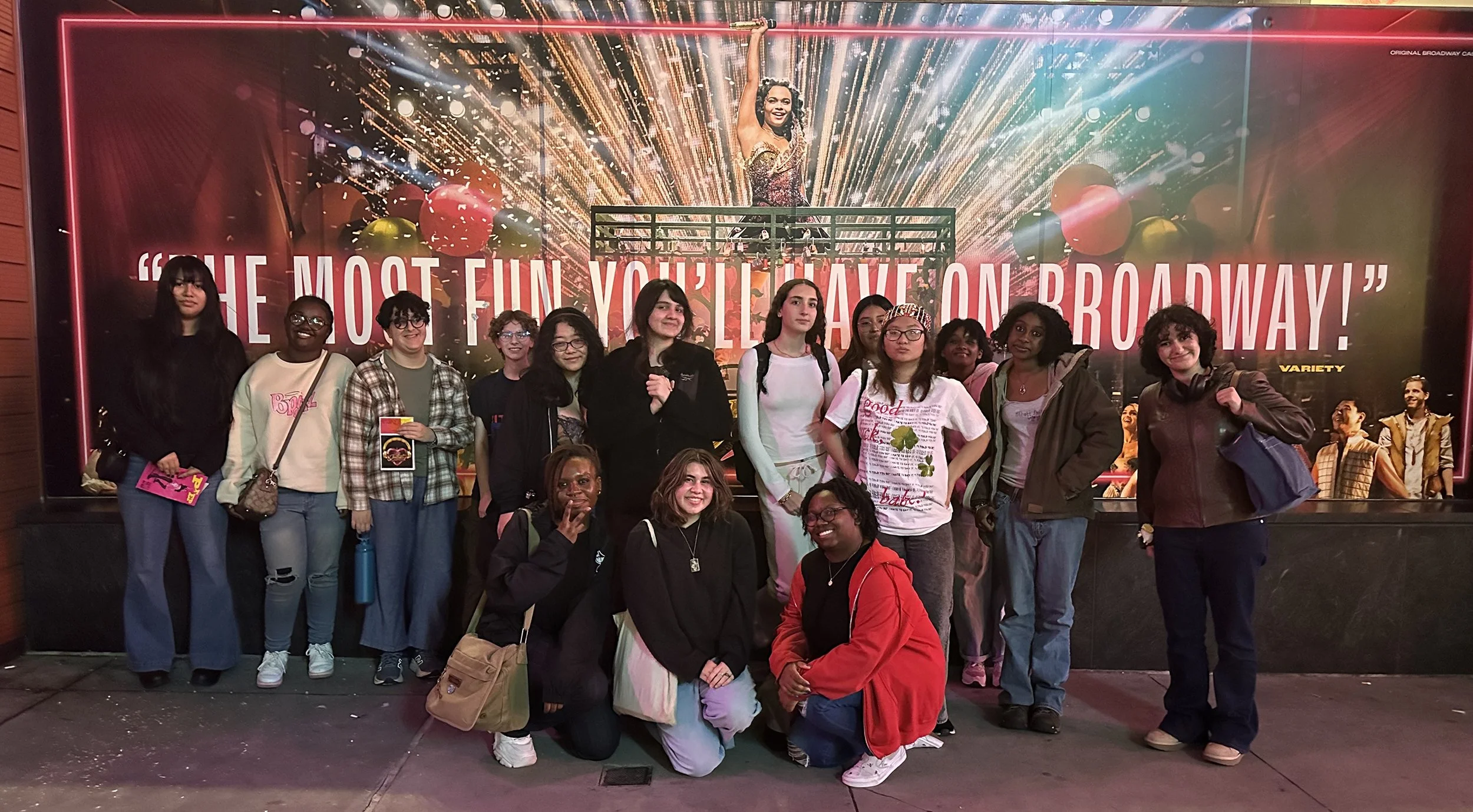 Group of diverse young women standing in front of Broadway theater poster, some smiling and holding items, others with hands in pockets, at night.