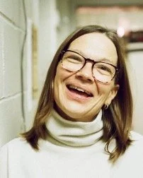 A woman with glasses and long hair smiling in a hallway.