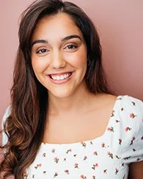 A young woman with long, dark hair smiling, wearing a white blouse with small floral patterns.