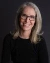 A woman with shoulder-length gray hair, glasses, and a black top, smiling at the camera.