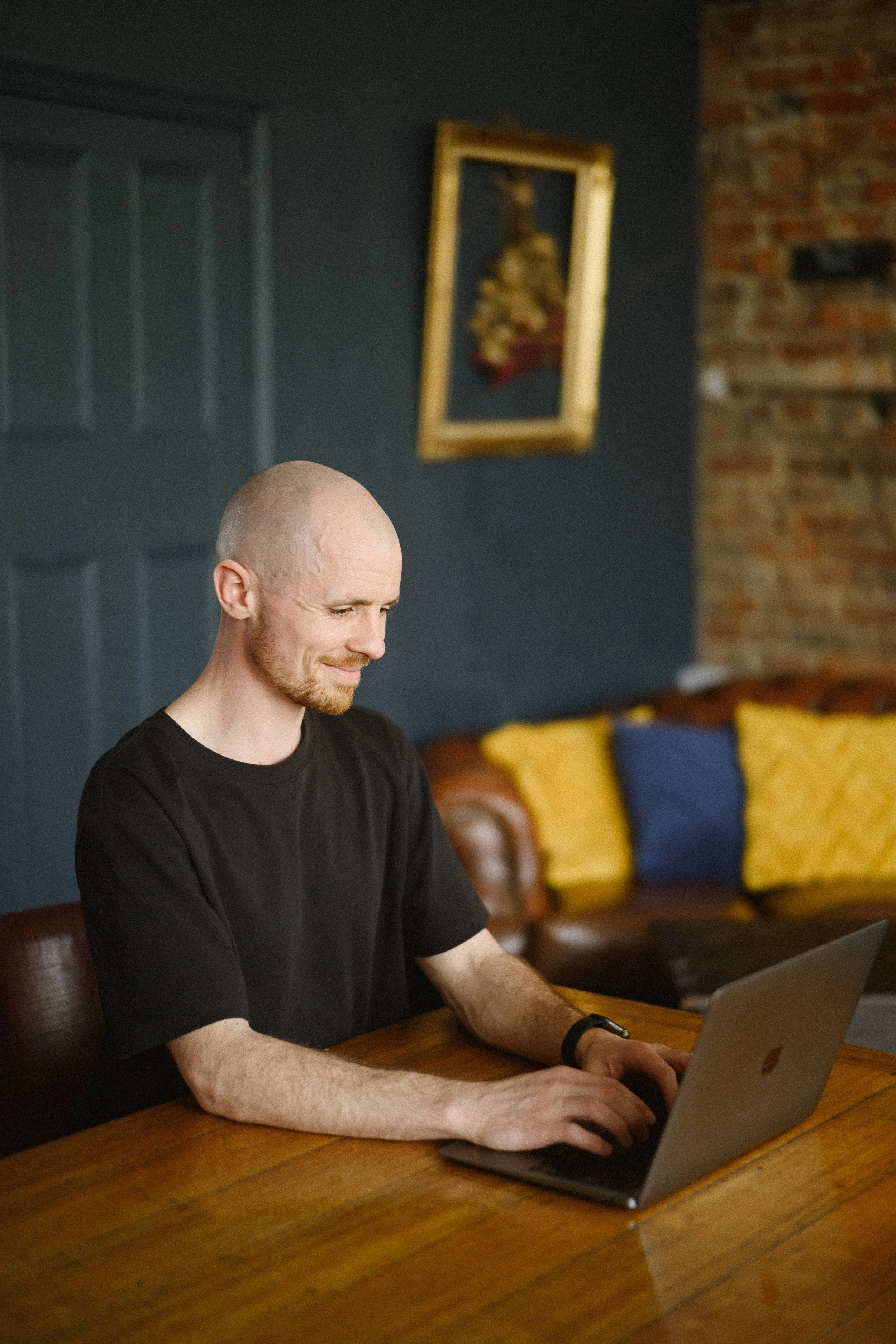 A man with a shaved head and beard sitting at a wooden table using a laptop in a cozy room with a dark wall, yellow and blue pillows, a leather couch, and artwork on the wall.