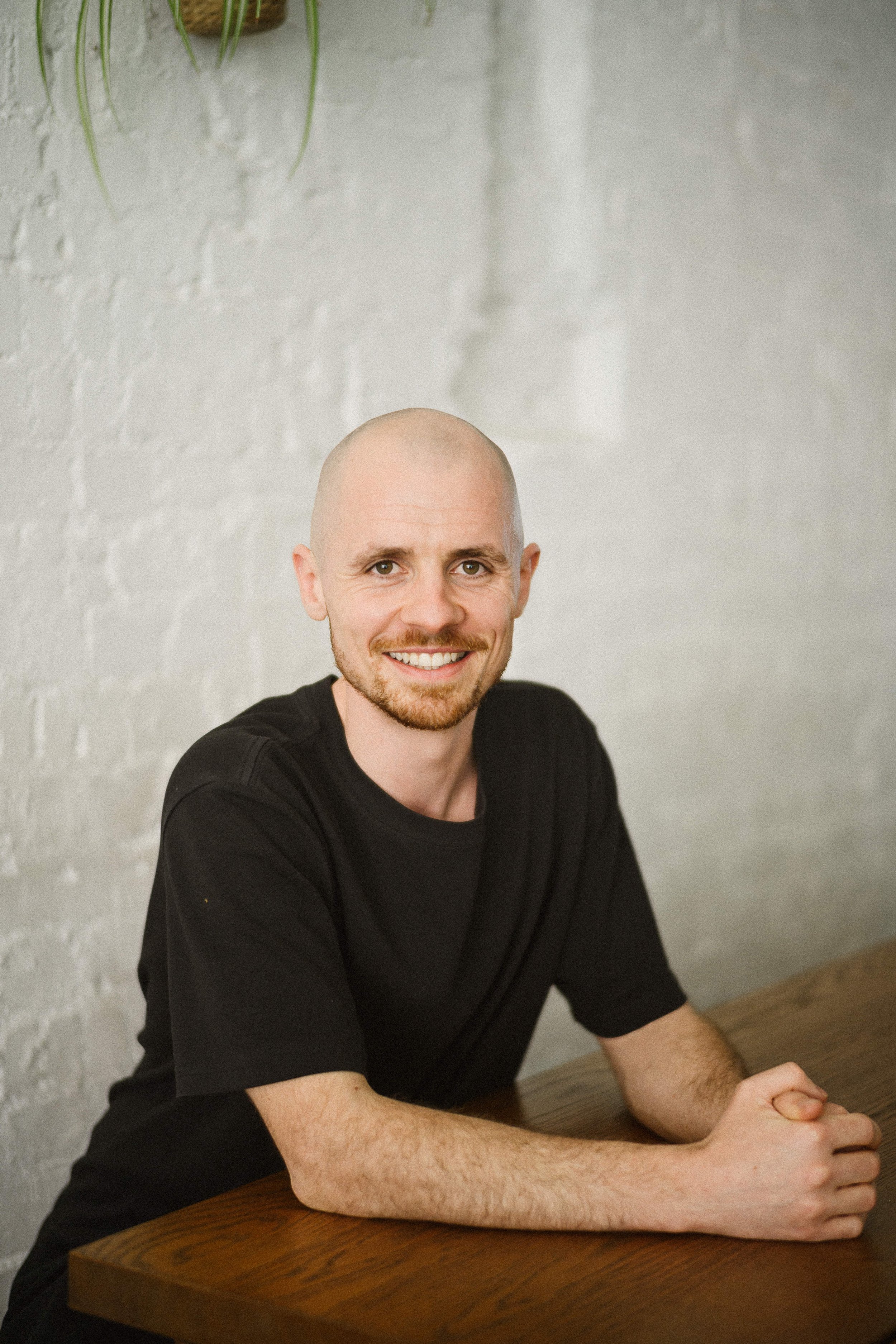 A smiling man with a shaved head and goatee sitting at a wooden table against a white brick wall.