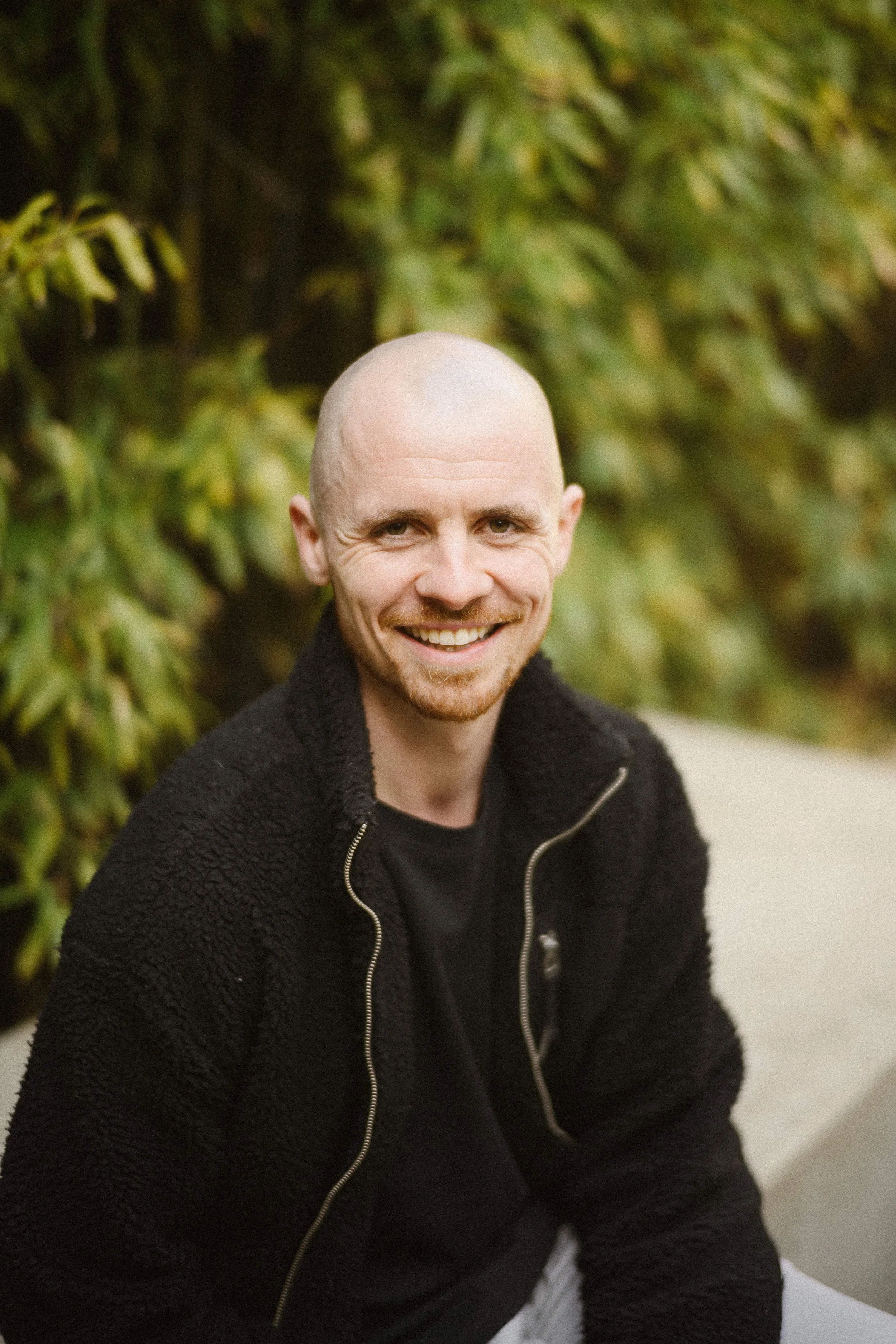 A smiling man with a bald head and a beard, wearing a black fleece jacket, sitting outdoors in front of green foliage.