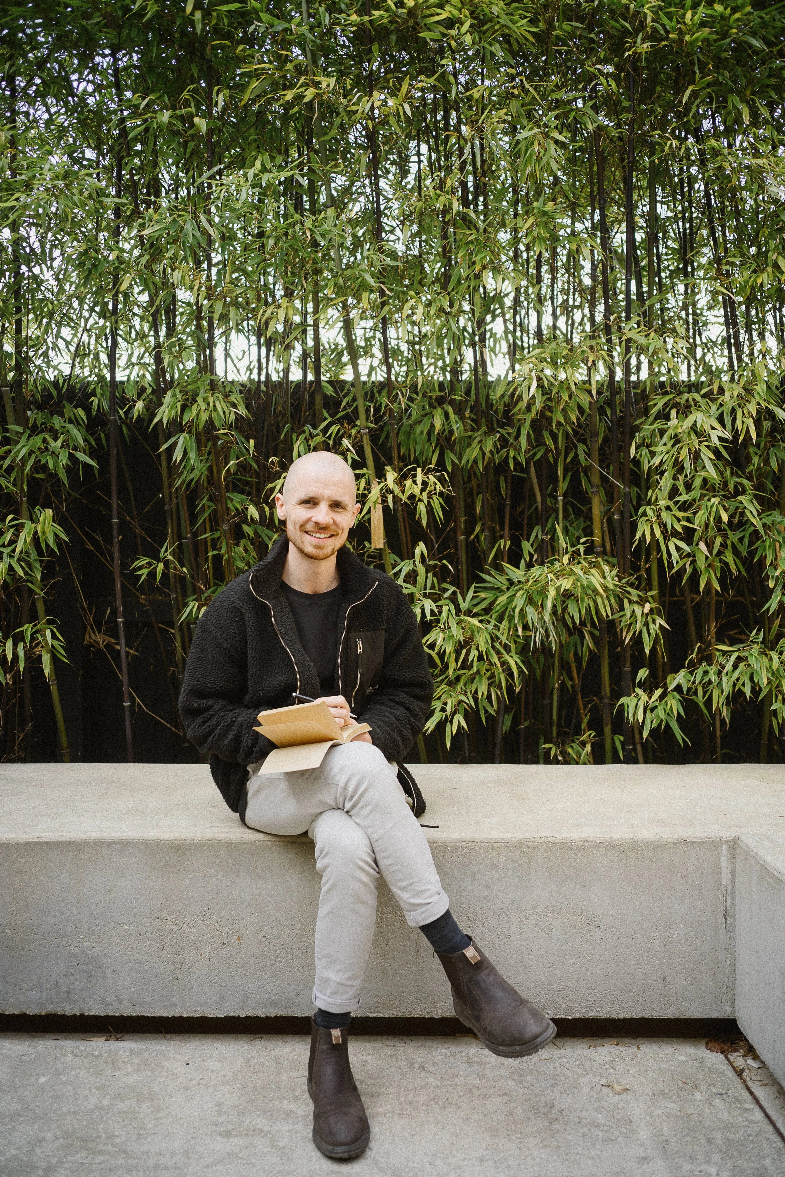 A man sitting on a concrete bench outdoors, smiling, holding a notebook and pen, with a background of tall green bamboo plants.