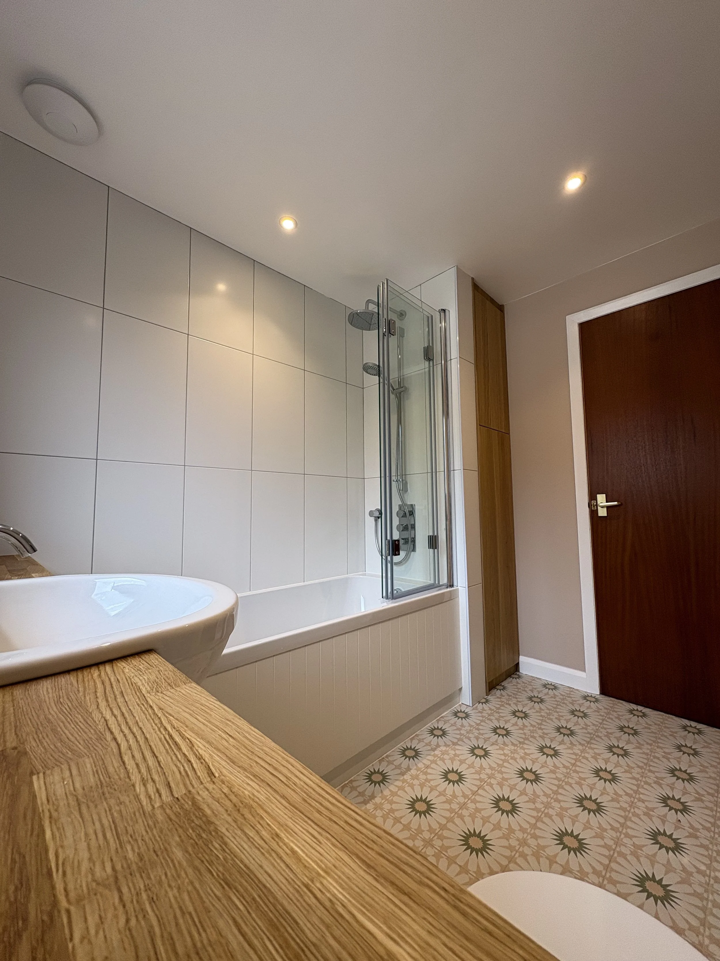 Modern bathroom with white tiled wall, glass shower enclosure, wooden cabinet, patterned tile floor, and wooden door.