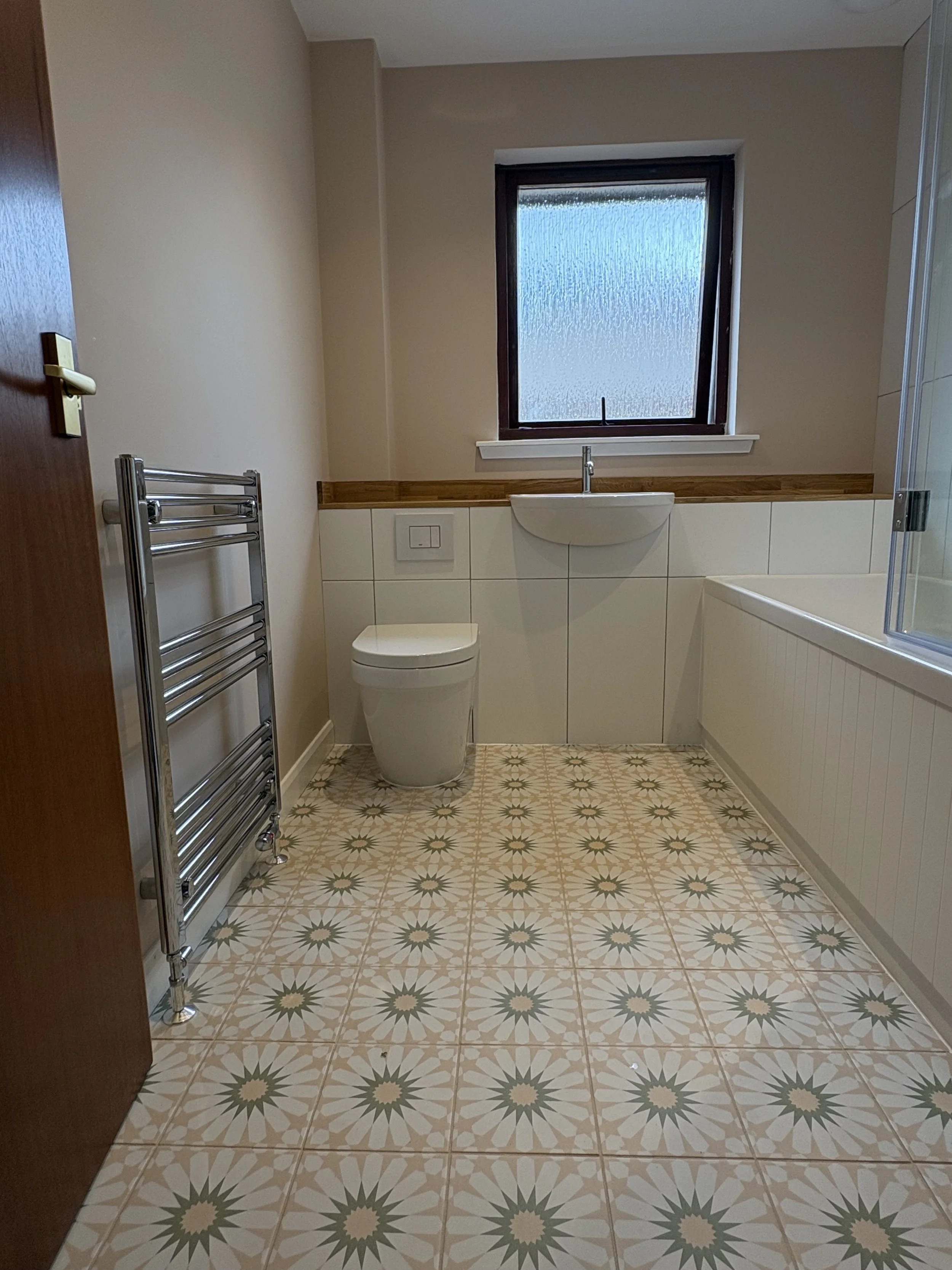 A bathroom with beige walls, a frosted window, a white toilet, a wooden ledge with a sink, white tiled walls, a towel rack, and patterned floor tiles.