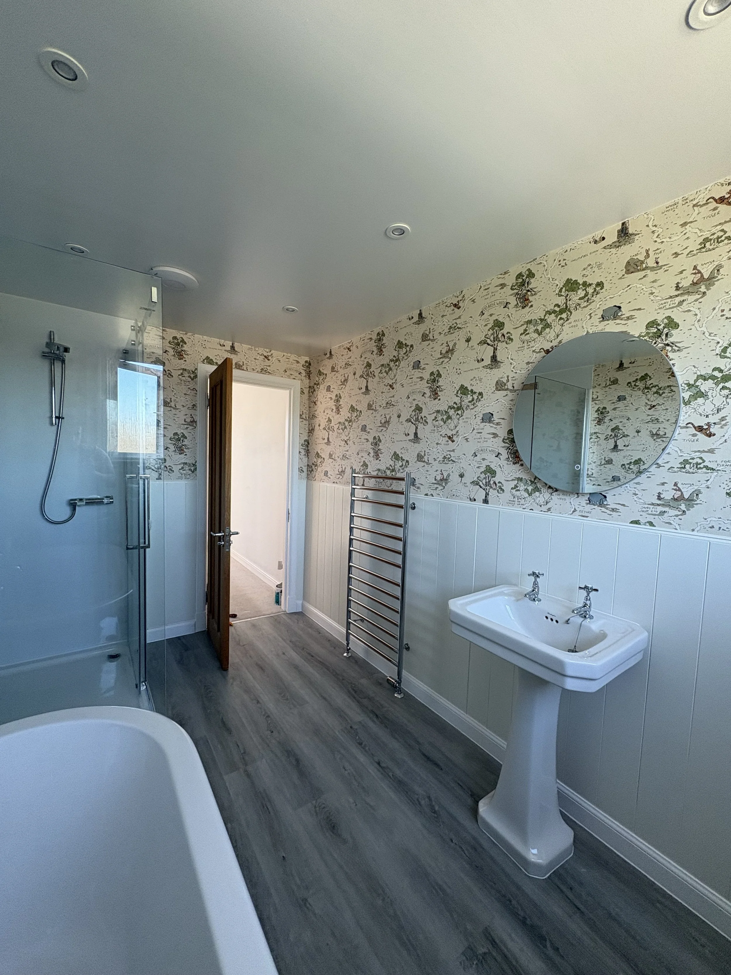 A bathroom with a white pedestal sink, a round mirror, a heated towel rail, and a wallpaper with a woodland scene featuring animals and trees.