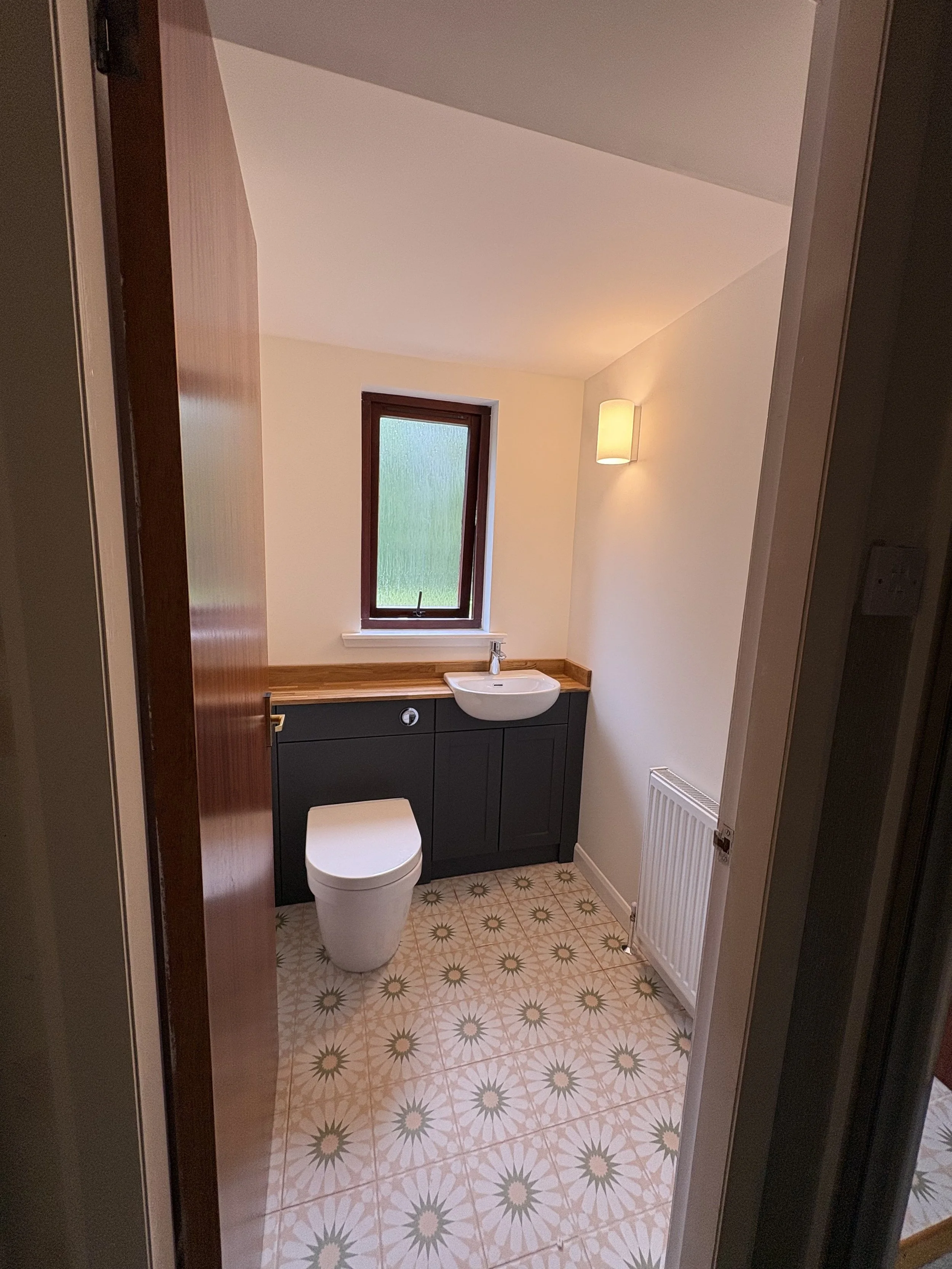 Small bathroom with a white toilet, a dark cabinet, a small sink, a window, a wall sconce, a radiator, and patterned floor tiles.