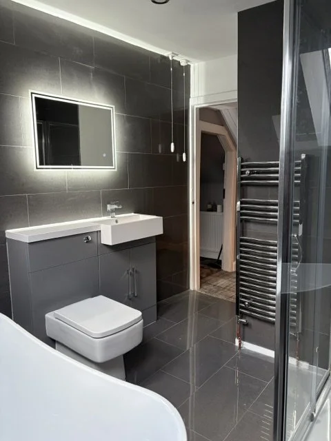 Modern bathroom with dark tiled walls and floor, illuminated mirror, white sink, gray cabinet, wall-mounted toilet, towel radiator, and glass shower enclosure.