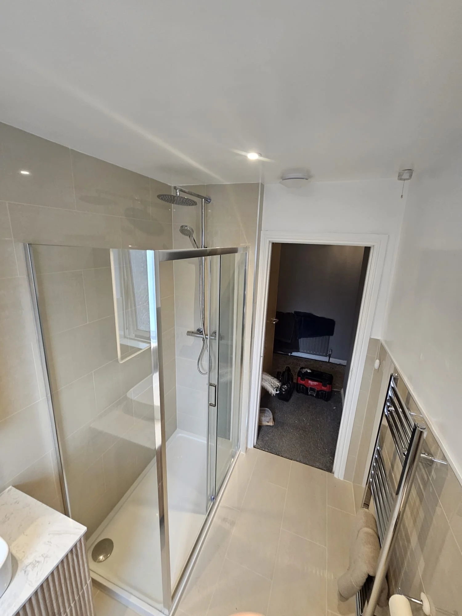 View of a bathroom with a glass shower enclosure, beige tiled walls, a white ceiling with recessed lights, a door open to a laundry room with tools and boxes, and a towel radiator on the right.