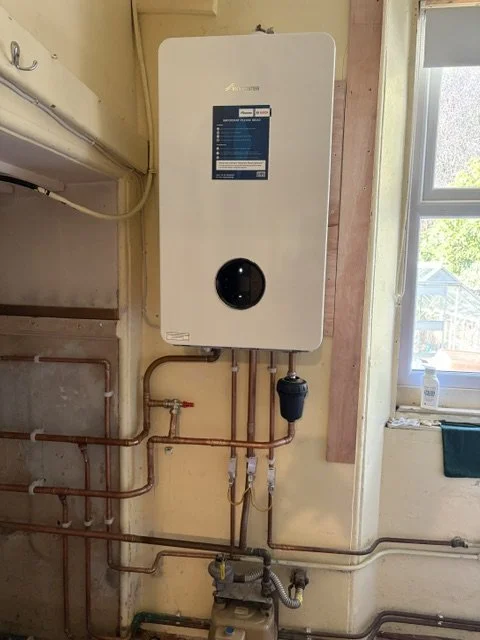 Wall-mounted tankless water heater with pipes and a small filter or pump underneath, next to a window.