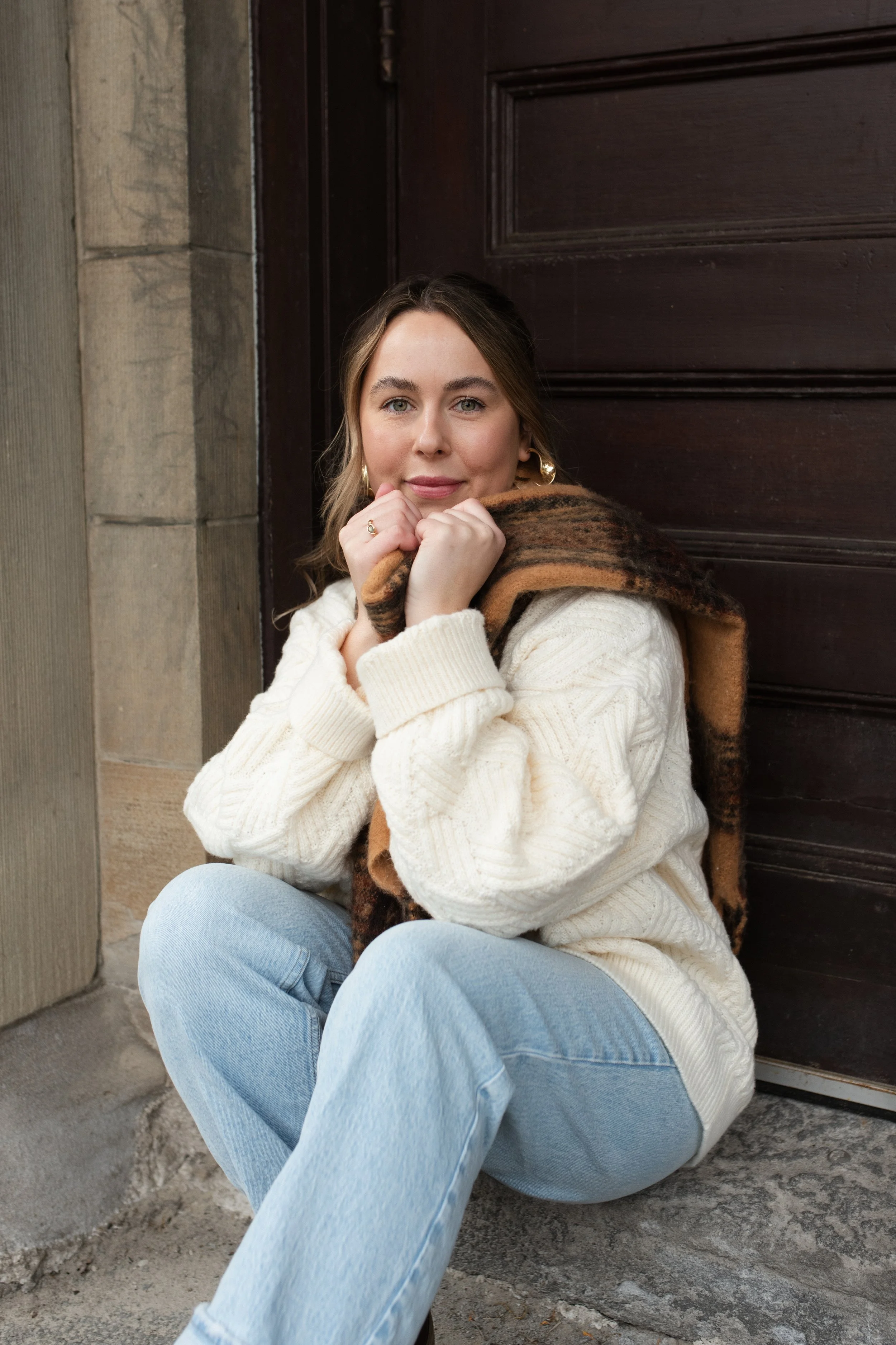 A woman sitting on the ground outside near a dark wooden door, wearing a white knitted sweater and light blue jeans, holding a brown and black scarf close to her face, with a gentle smile.
