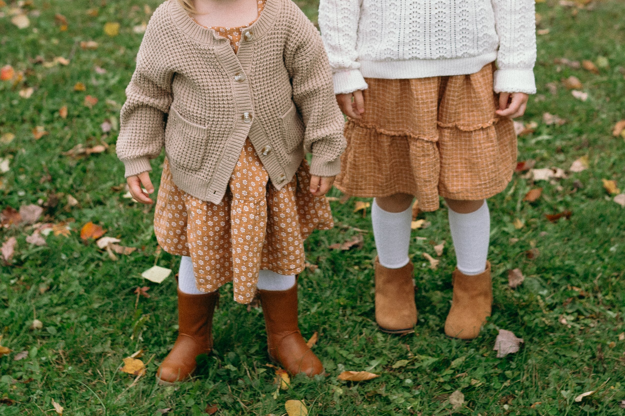 Two children standing outdoors on grass with fallen leaves, wearing brown boots, white leggings, brown skirts, and warm sweaters.