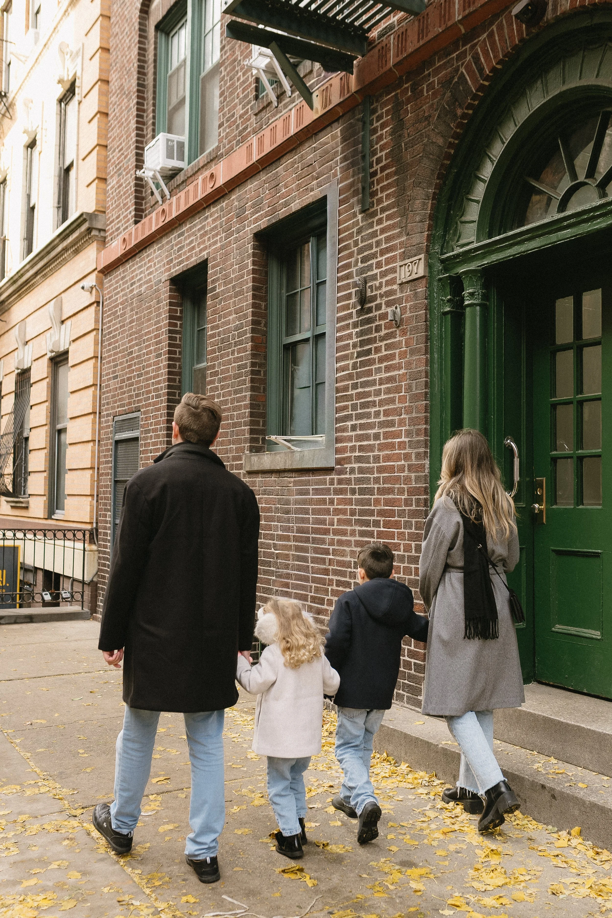 A family of four walking along a city sidewalk with fallen yellow leaves. They are all dressed warmly, with the mother and children wearing coats. They are near a brick building with green doors and window frames.