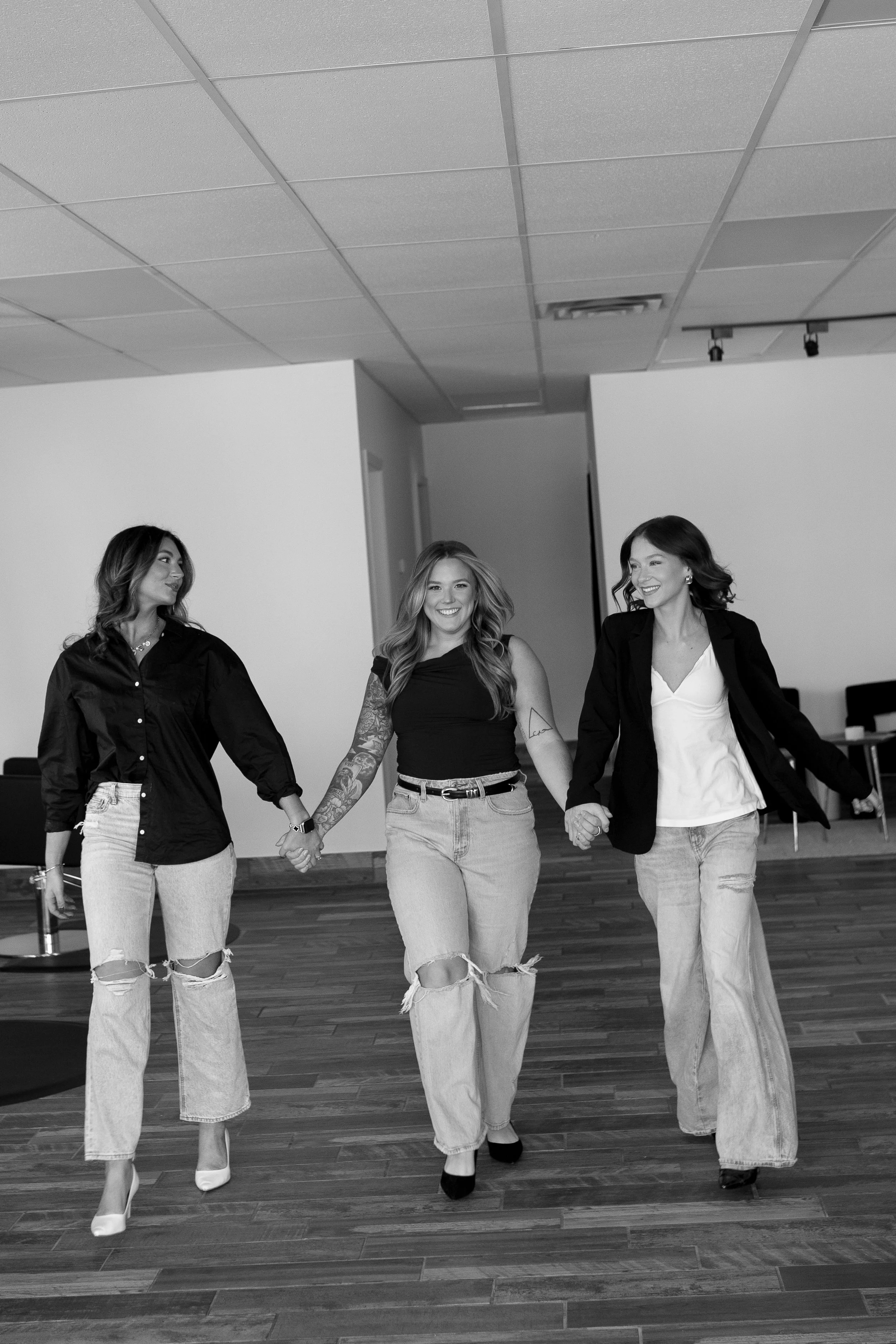 Three women holding hands and walking in a room with wooden flooring, smiling and looking at each other.