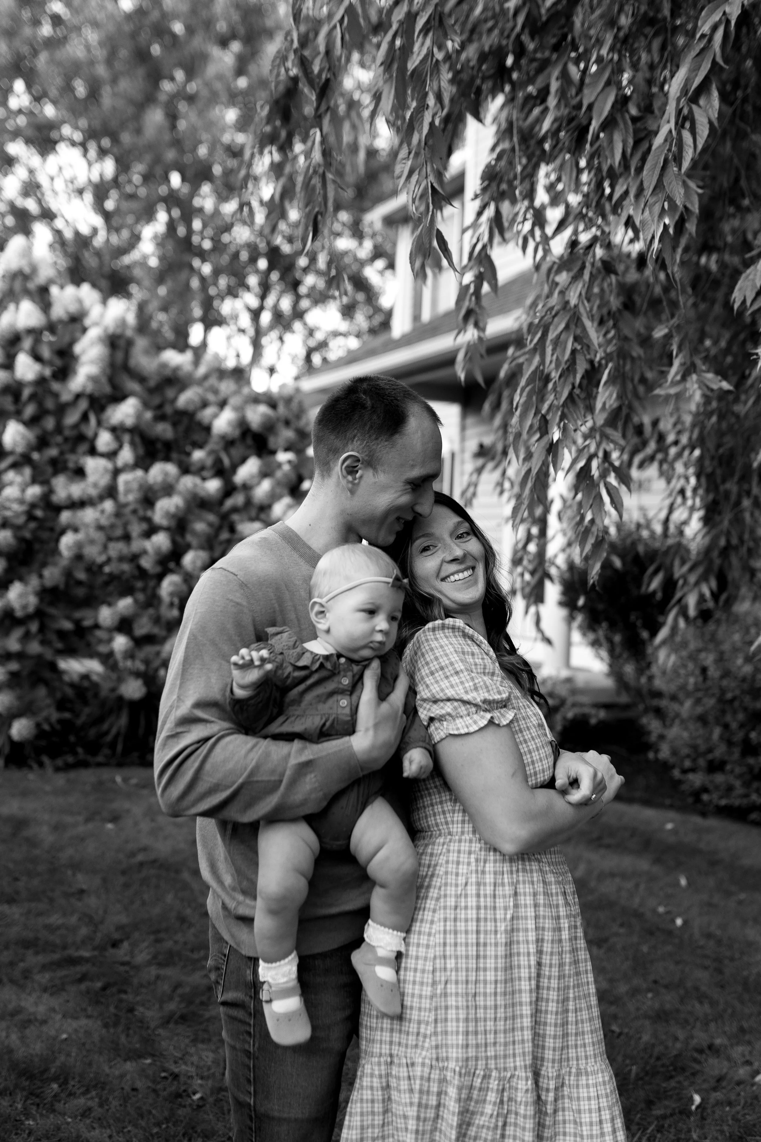 A black and white photo of a happy family of three outdoors in a garden. The father holds a baby girl, and the mother stands beside them, smiling.