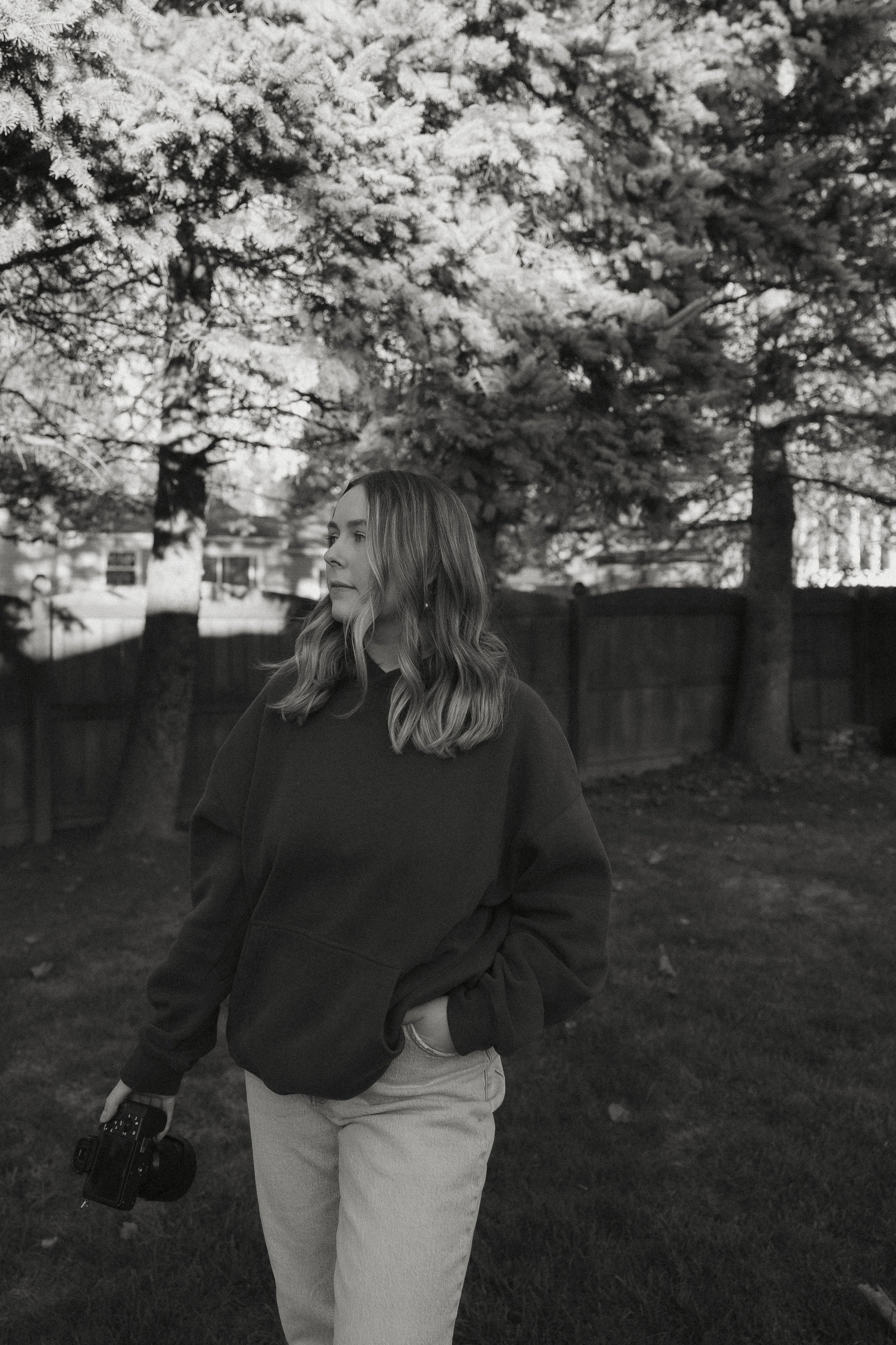 A young woman with long wavy hair, wearing a dark hoodie and light pants, stands outdoors in a yard with trees and a wooden fence in the background. She is holding a camera in her right hand and looks to the side.