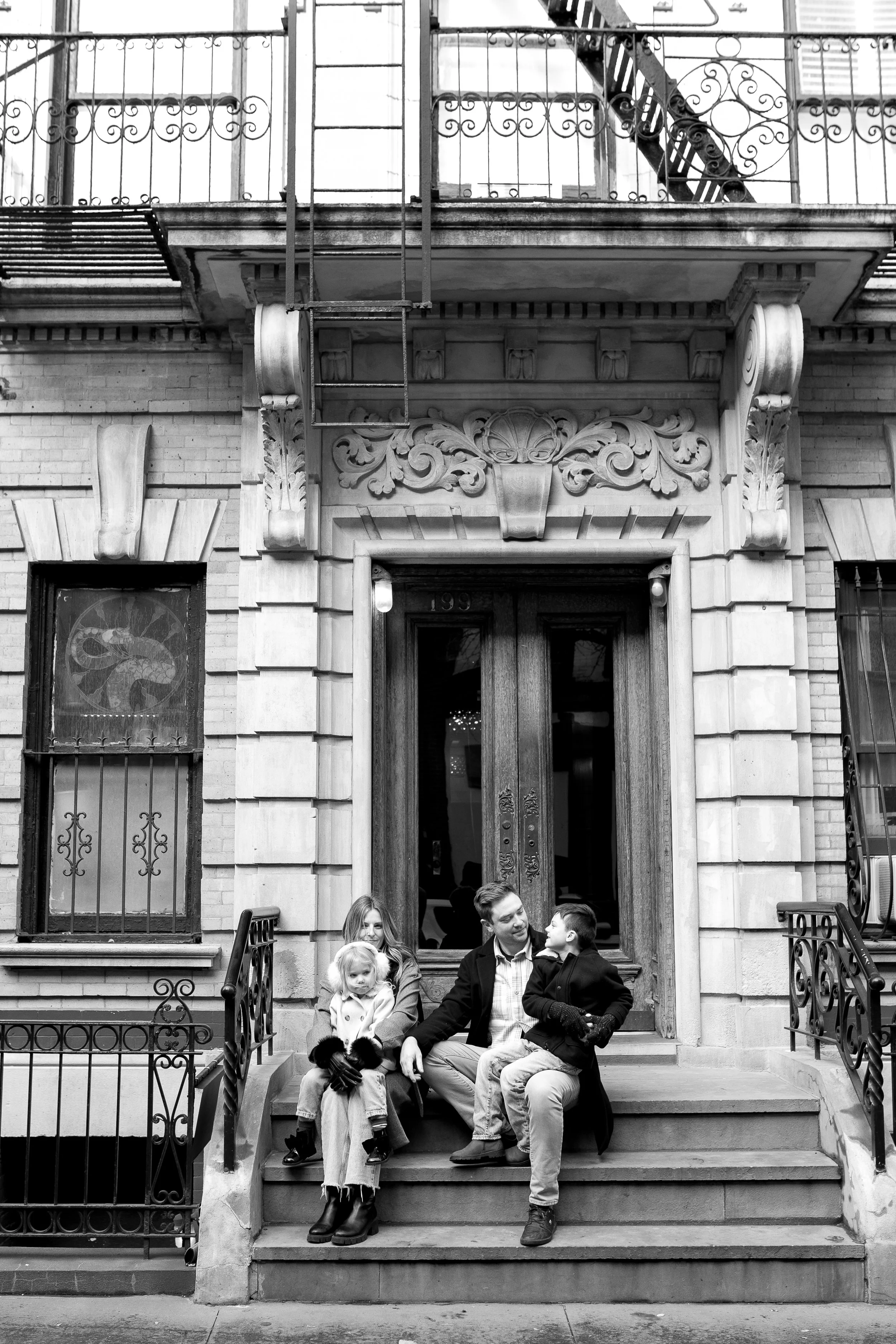A family sitting on the steps of a city brownstone. The father is talking to a boy, and the mother and a girl are seated nearby. The building has ornate architectural details and fire escapes above.