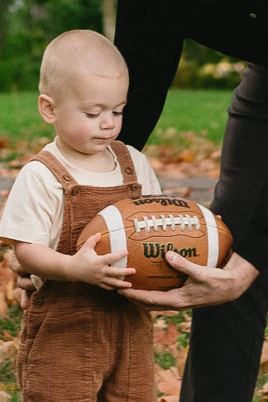 Young child with a shaved head, wearing brown overalls and a white shirt, holding a football while an adult helps support it outdoors in a park.