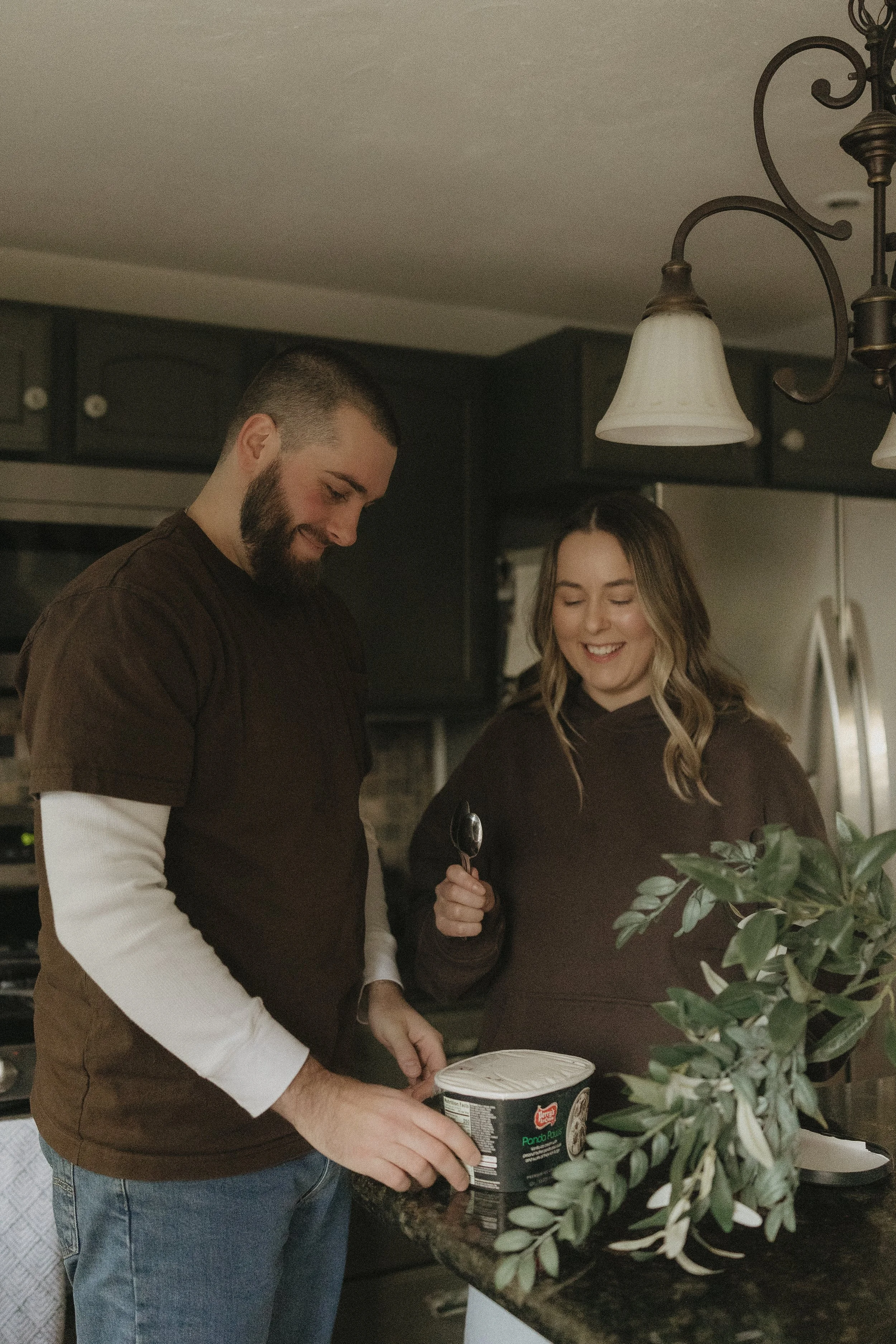 A smiling man and woman in a kitchen, looking at ice cream container on the counter, with greenery nearby.