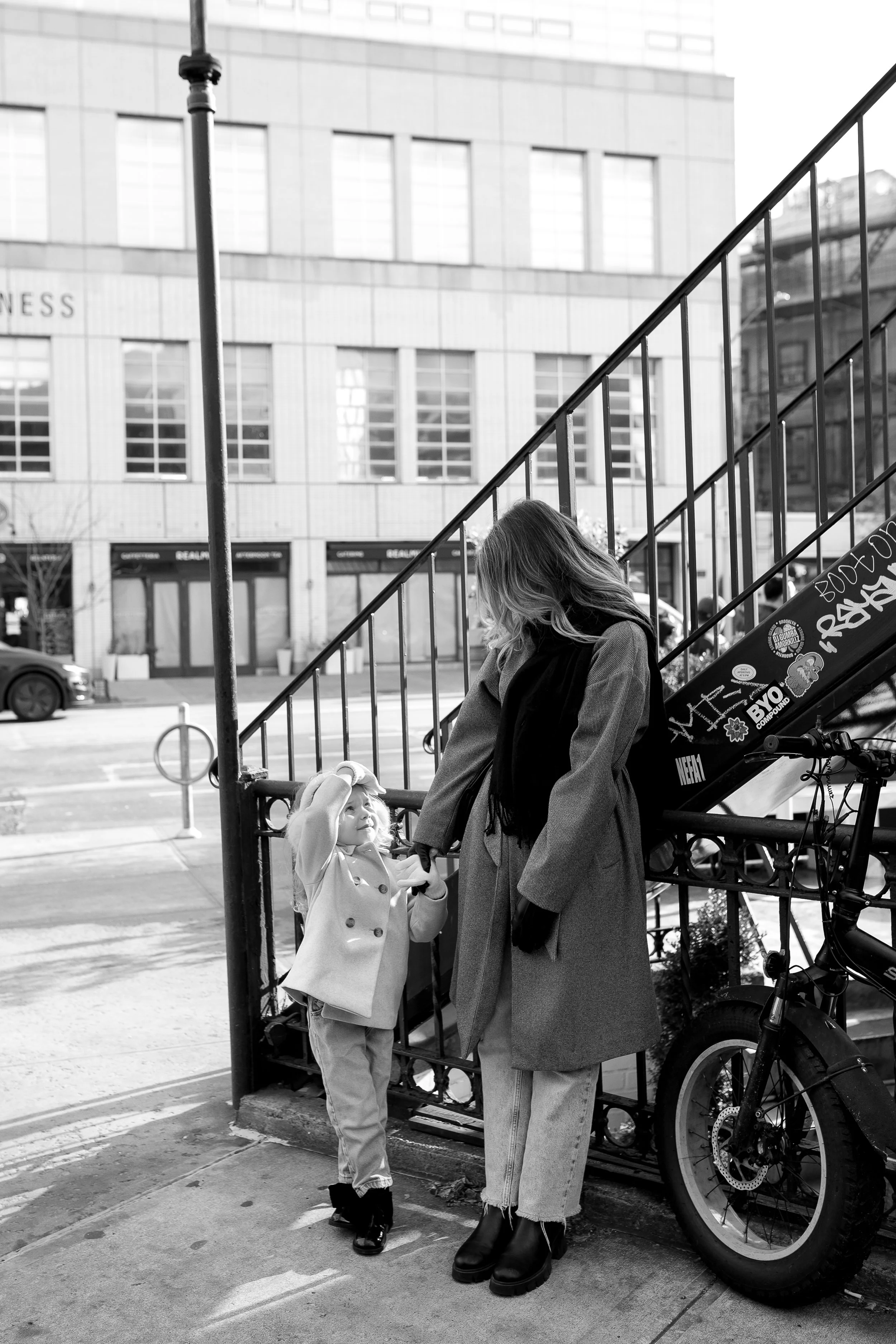 A woman and a young girl standing outside in an urban area, interacting near a staircase and a bicycle, with a city building in the background.